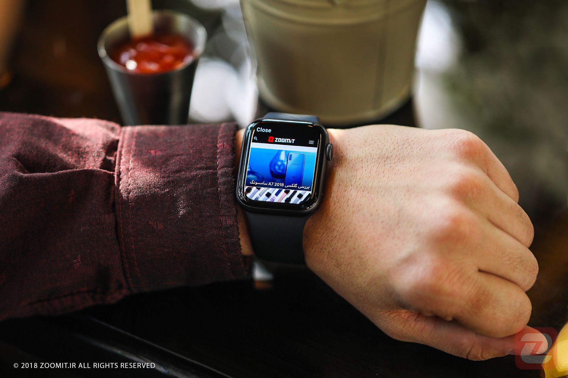 بررسی اپل واچ سری ۴ / Apple watch series 4 در دست چپ روی میز کافه درحال نمایش سایت زومیت