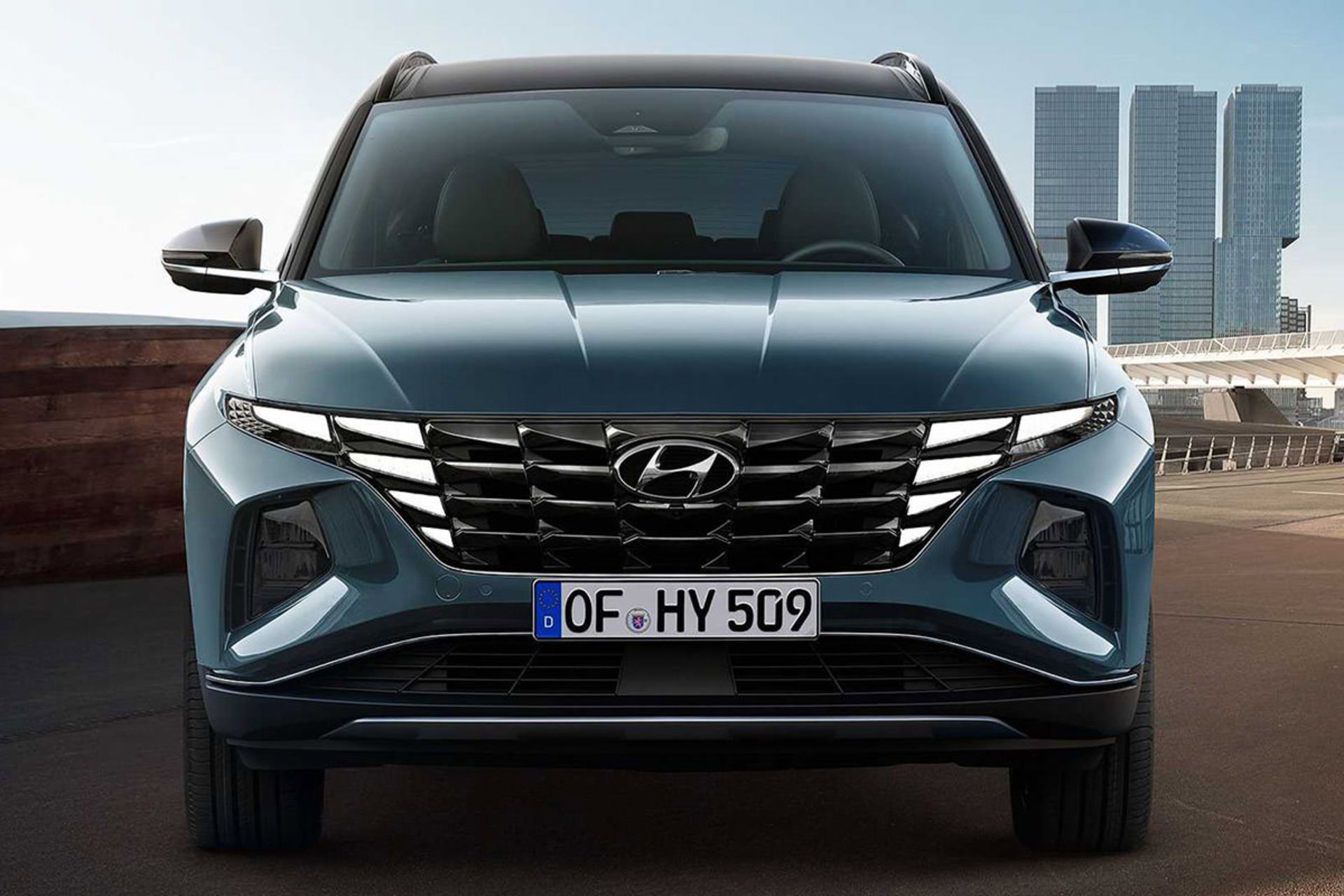 نمای جلو خودرو کراس اور هیوندای توسان 2022 / 2022 Hyundai Tucson crossover با رنگ آبی در کنار ساختمان و آسمان آبی با ابر