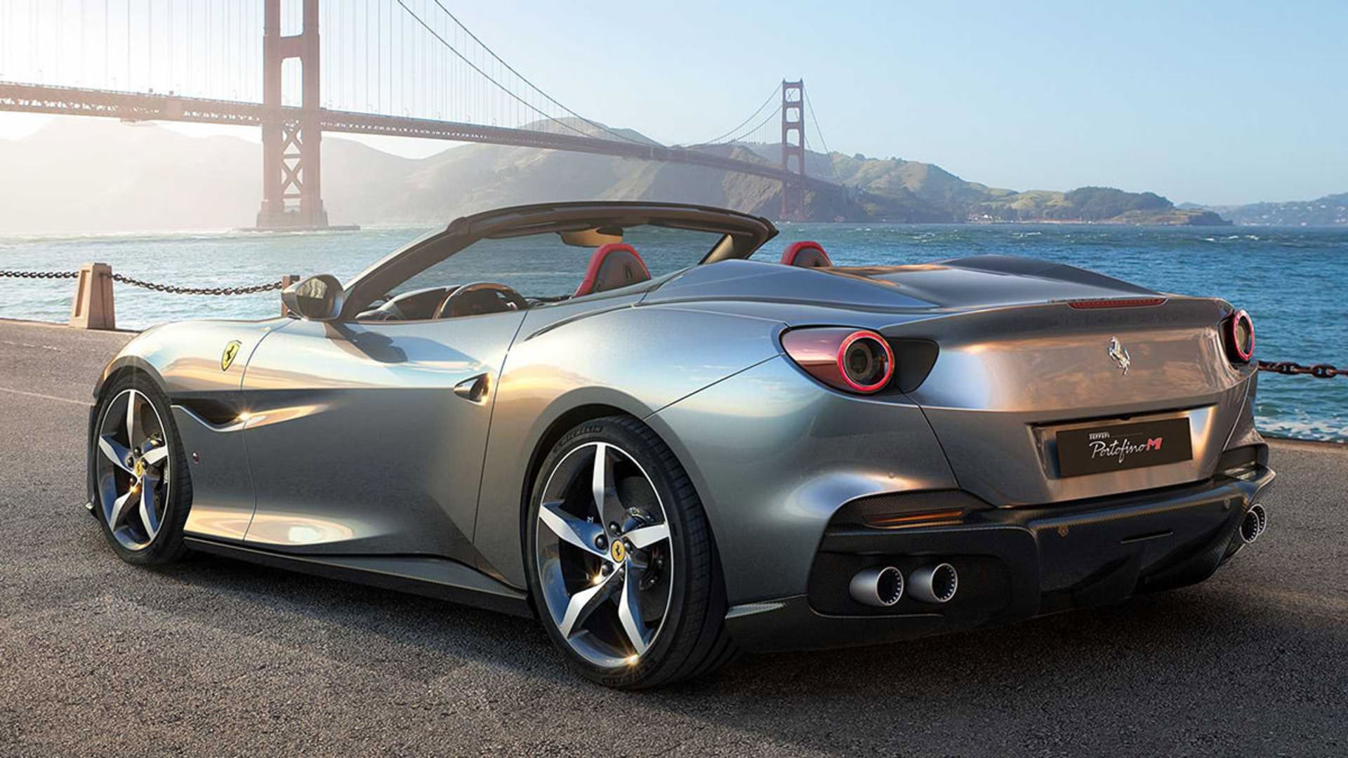 نمای عقب خودرو روباز فراری پورتوفینو ام / Ferrari Portofino M نقره ای رنگ در کنار دریا و پل معلق