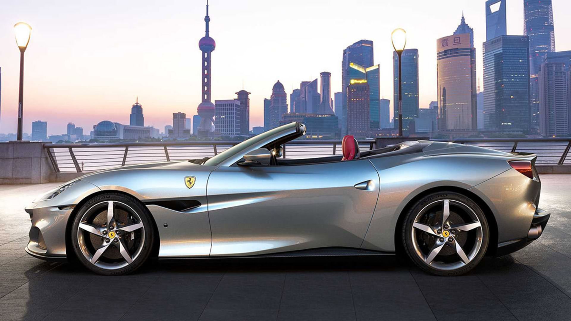 نمای جانبی خودرو روباز فراری پورتوفینو ام / Ferrari Portofino M نقره ای رنگ در کنار ساختمان و آسمان موقع غروب