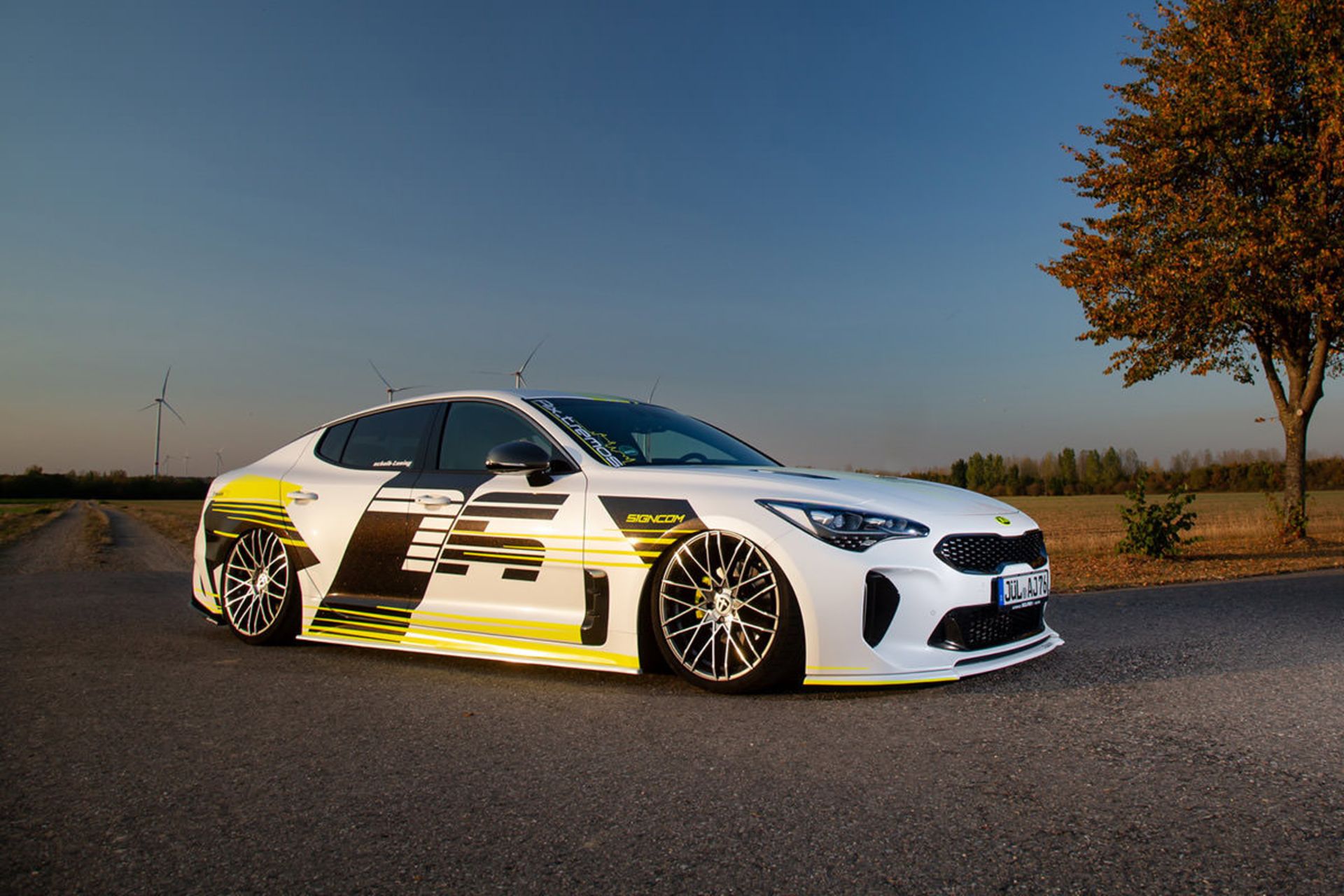 نمای جانبی کیا استینگر / Kia Stinger سفید رنگ با تزئینات نئون و منظره آسمان و درخت