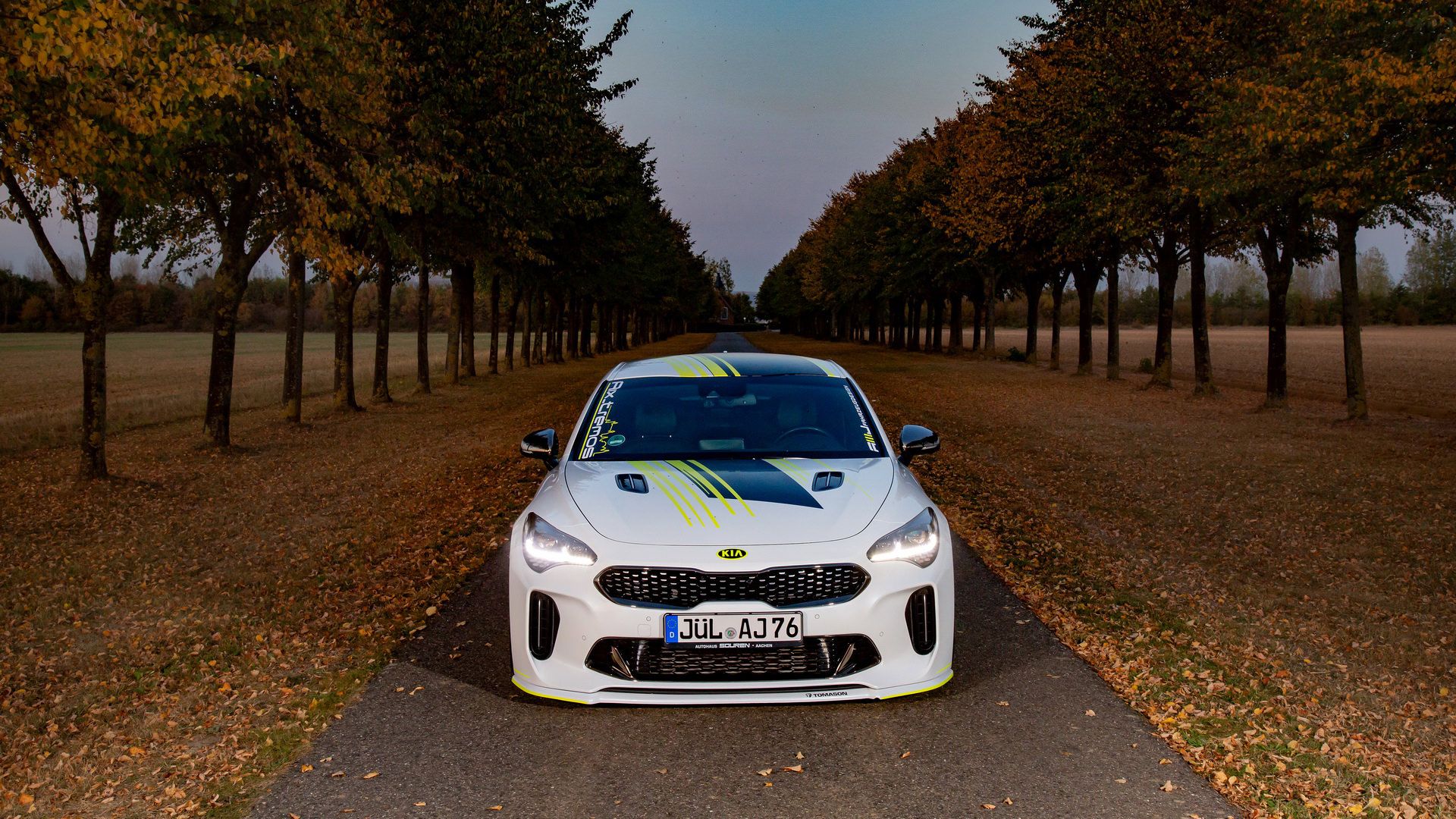 نمای جلو خودروی اسپرت کیا استینگر / Kia Stinger سفید رنگ با تزئینات نئون و منظره درختان 