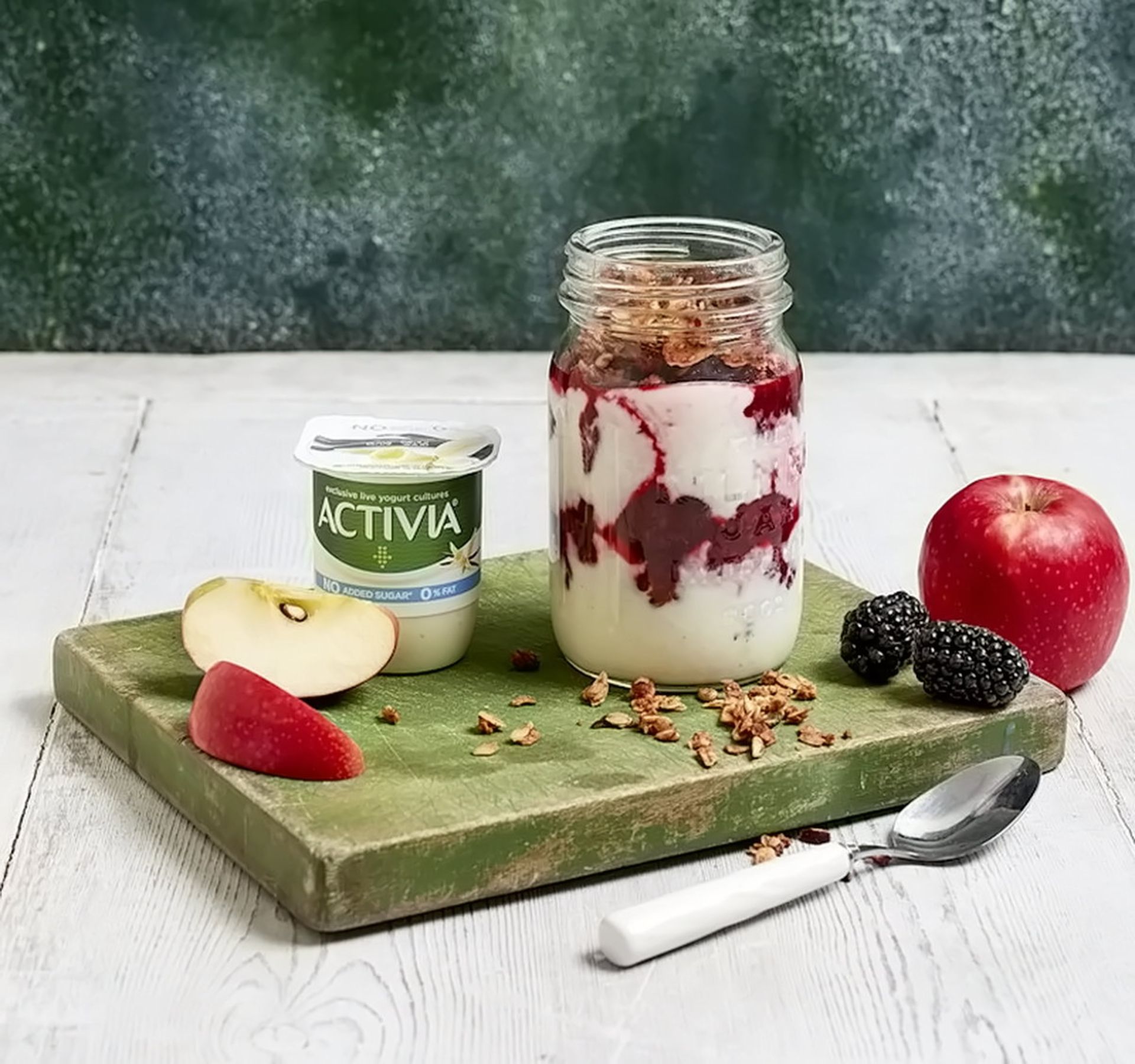 ماست اکتیویا برند دانون روی تخته به همراه برش سیب، چند توت سیاه و یک شیشه دسر