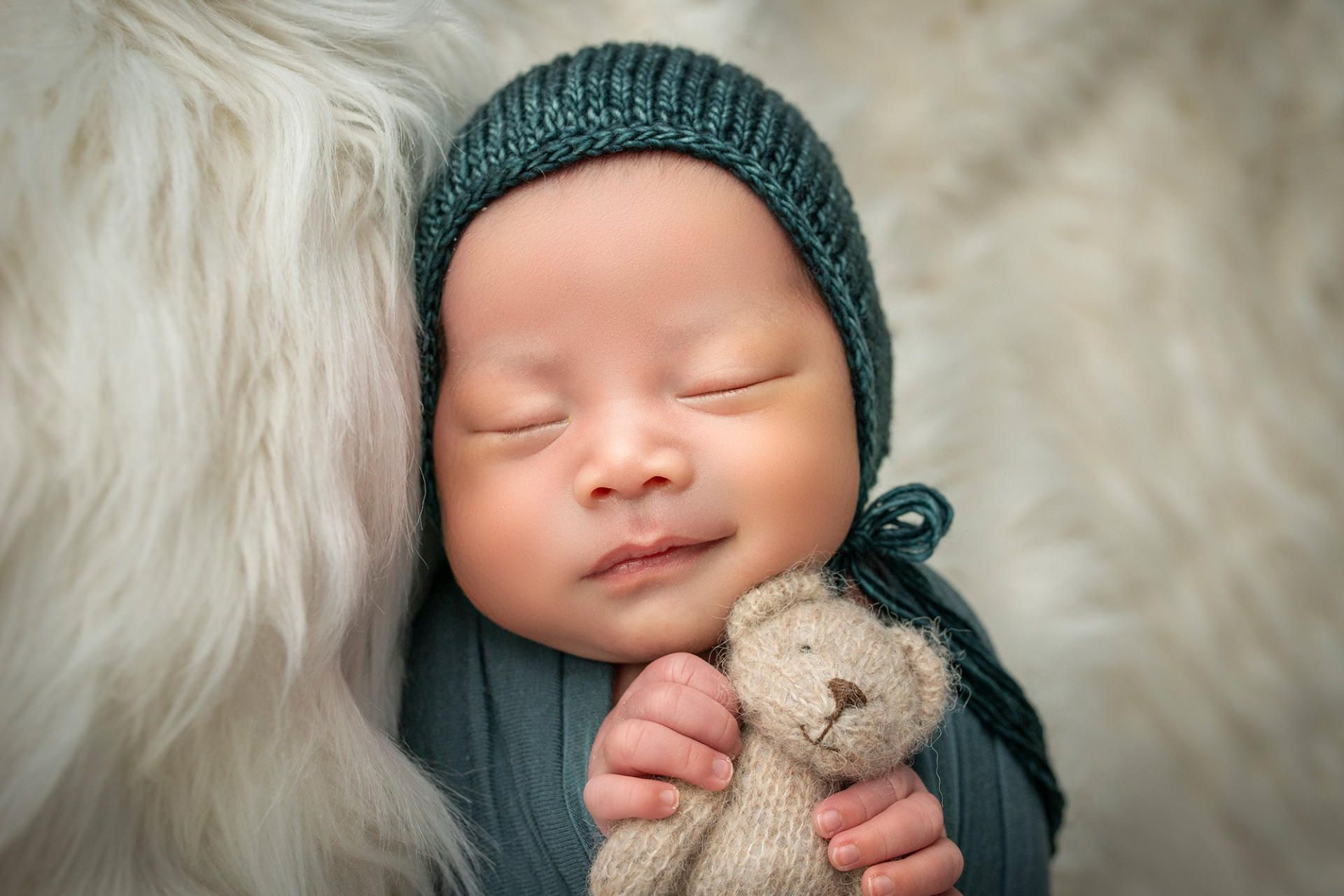 پسربچه نوزاد با لباس بافت خاکستری عروسک خرس را بغل کرده و خوابیده است