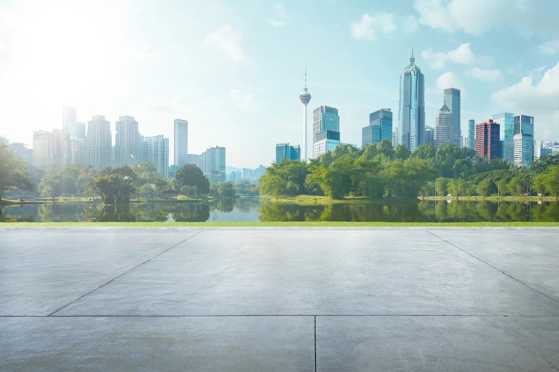 نمای یک شهر مدرن با برج‌های بلند در پس‌زمینه، در کنار دریاچه و فضای سبز در پیش‌زمینه، زیر نور سفید و روشن خورشید