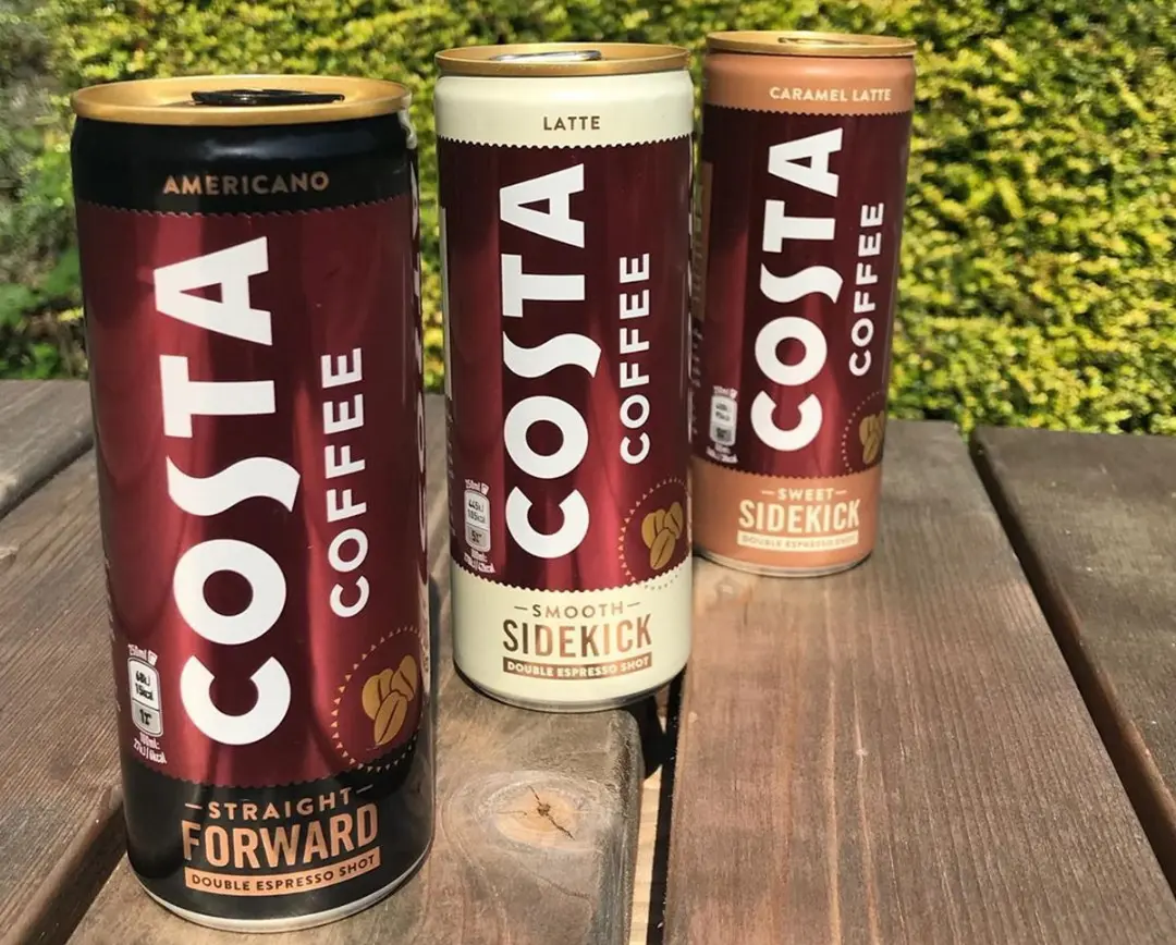 سه قوطی Costa Coffee روی نیمکت چوبی در فضای باز
