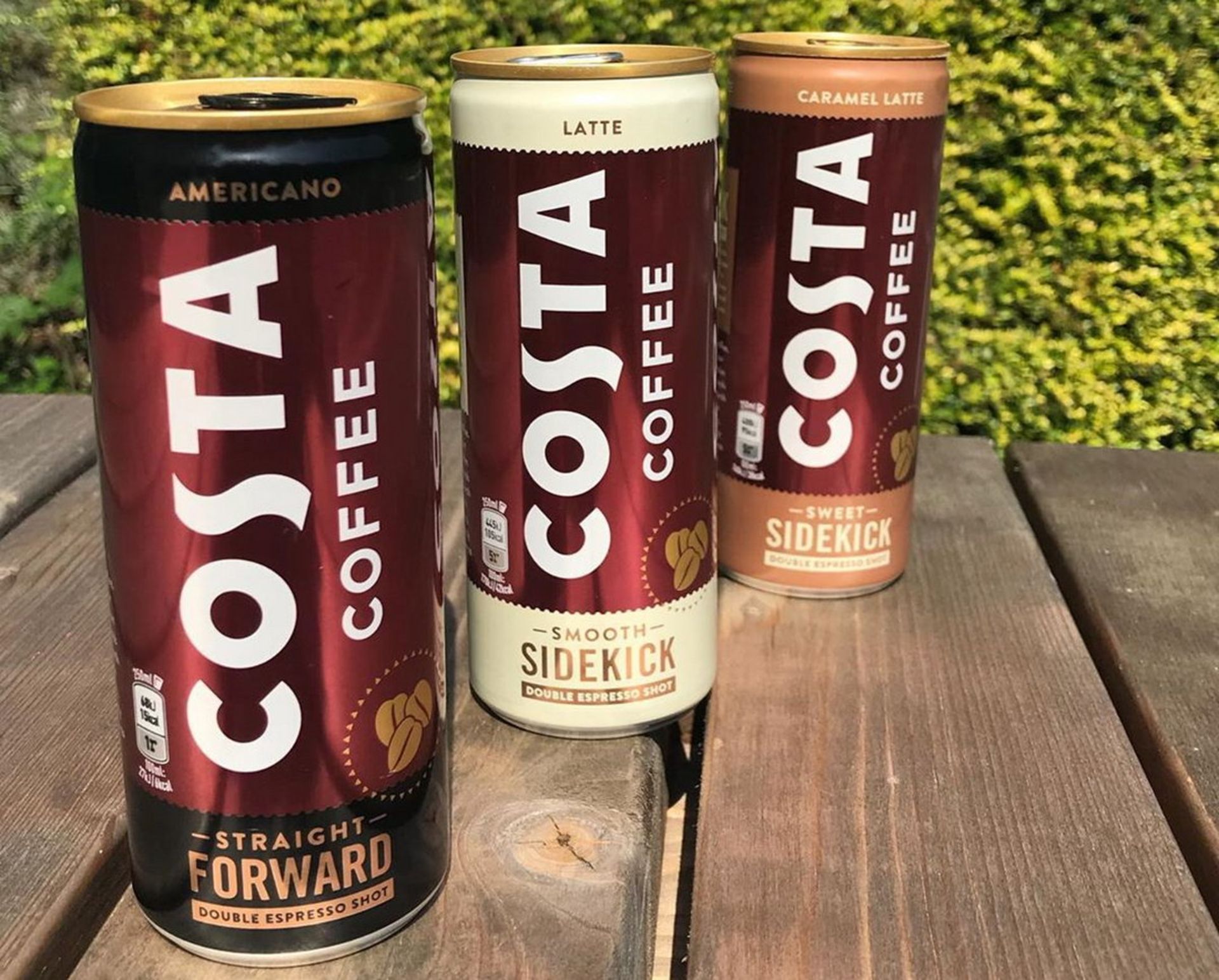 سه قوطی Costa Coffee روی نیمکت چوبی در فضای باز