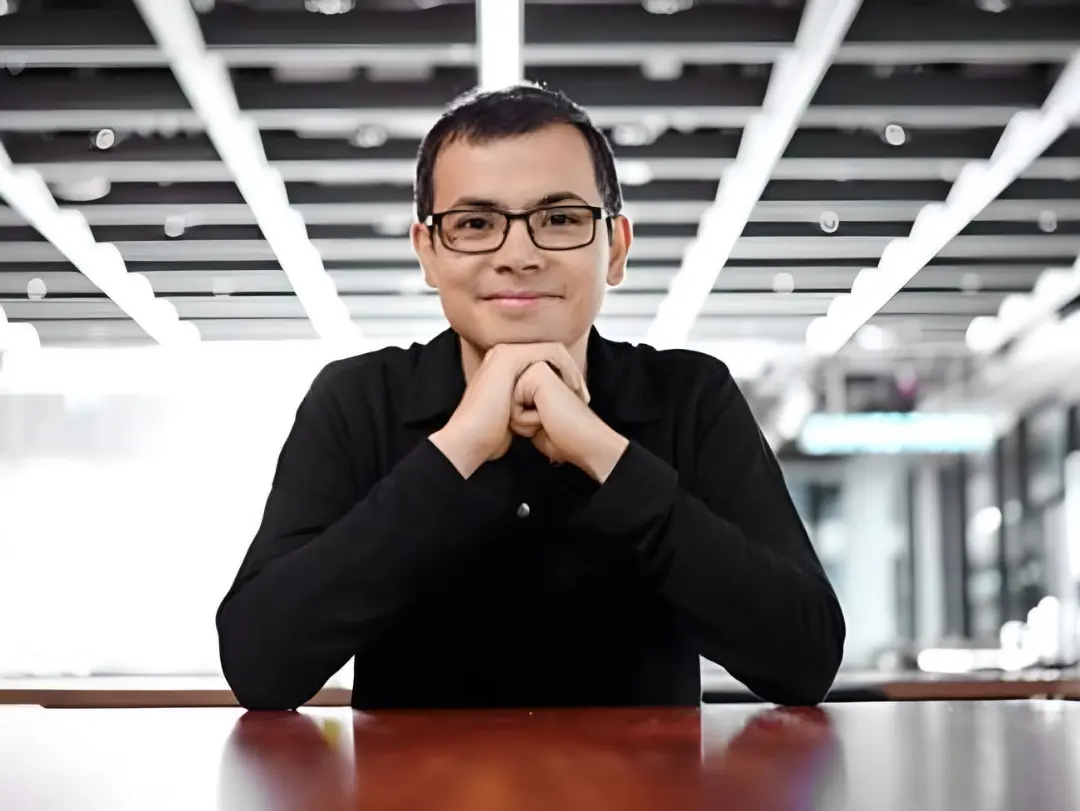 دمیس هاسابیس (Demis Hassabis) با عینک و لباس مشکی پشت میز نشسته است