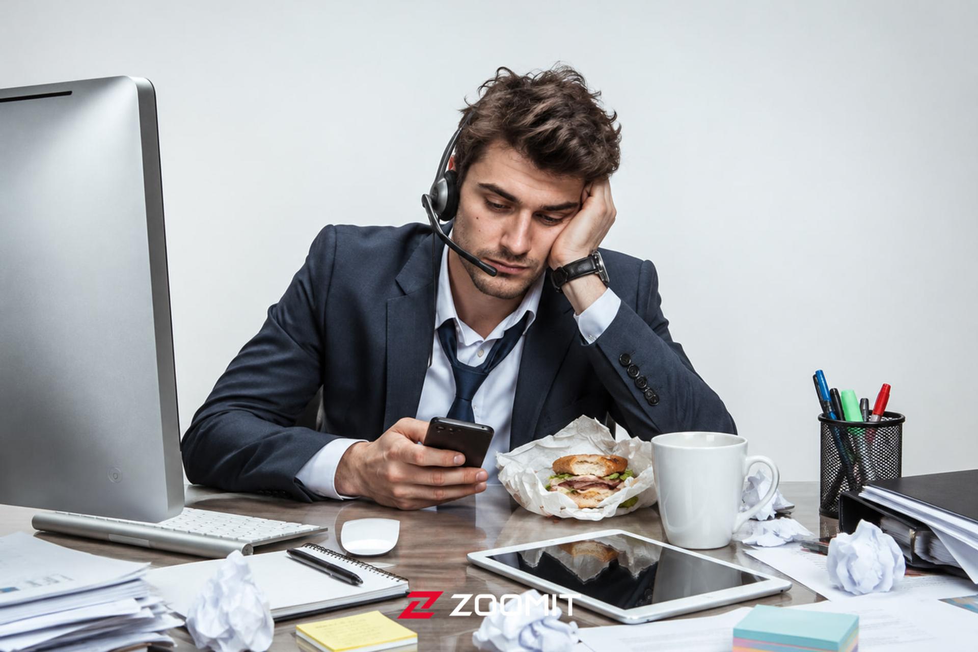 مردی خسته با هدست که در حال چک کردن گوشی هوشمند در میز کار است