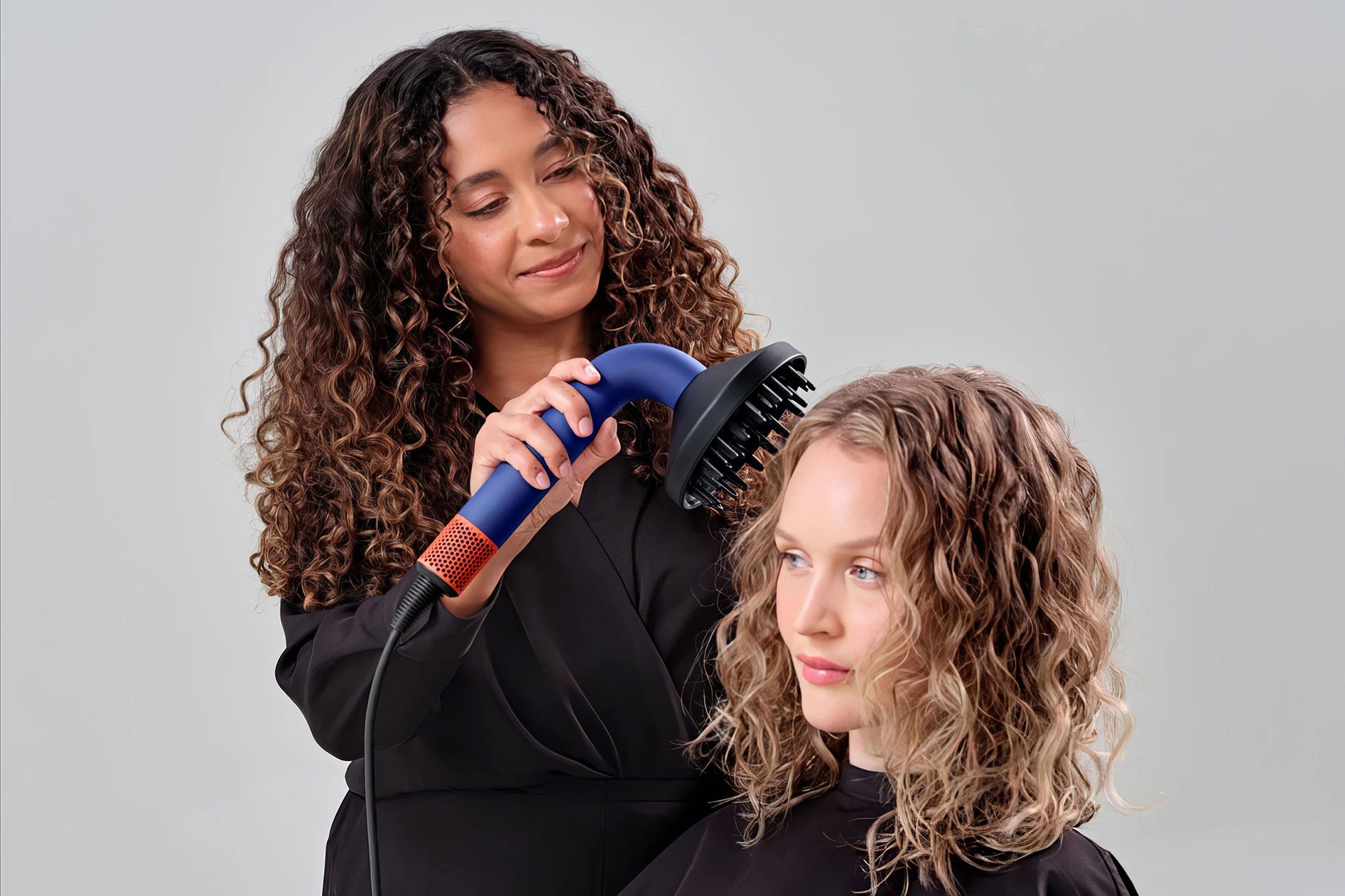 یک زن درحال استفاده از سشوار سوپرسونیک آر برای سشوار کشیدن سر یک زن که نشسته است