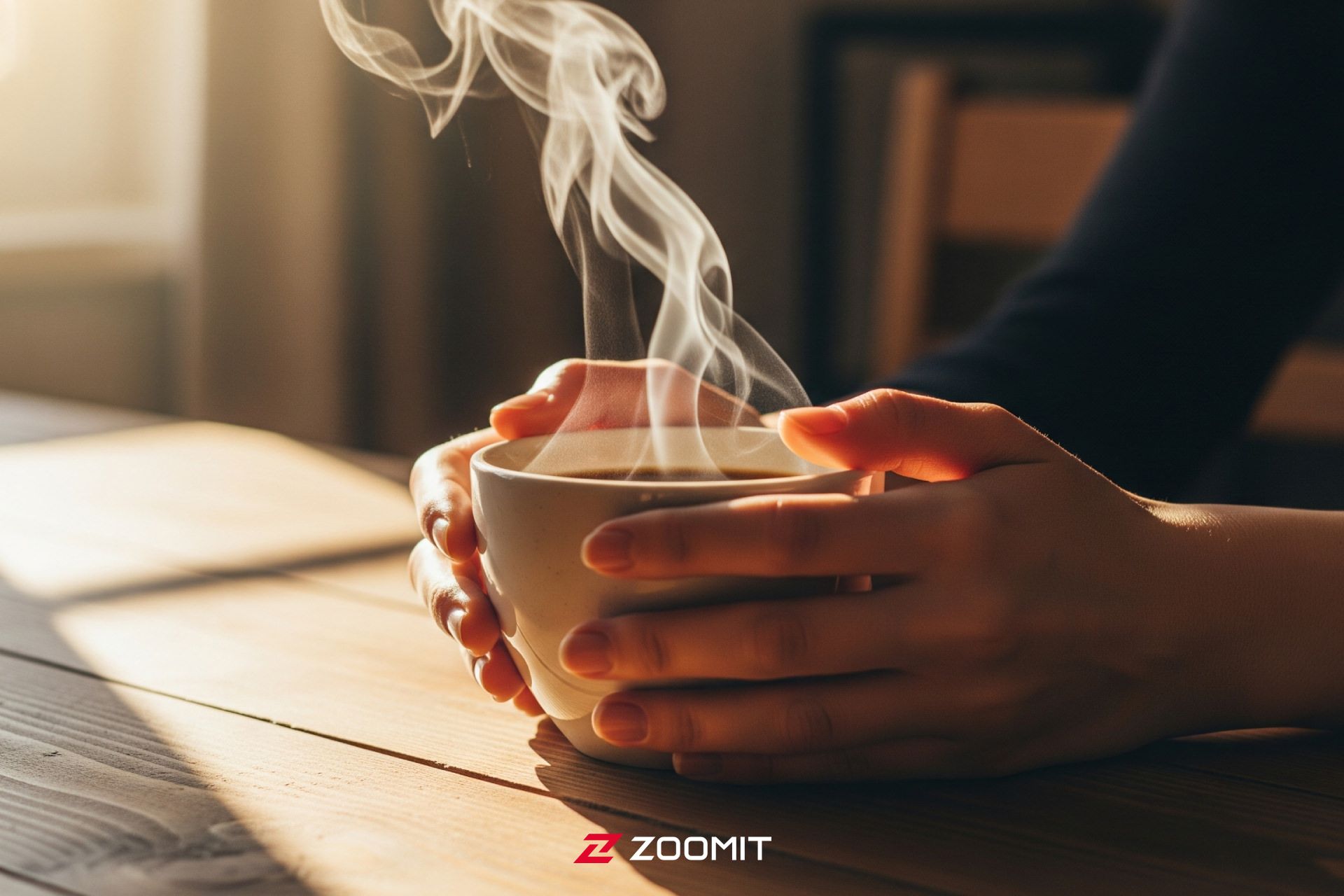 فنجان قهوه بخارآلود روی میز چوبی با نور ملایم صبحگاهی که از پنجره و دستان فردی که فنجان را در برگرفته است.