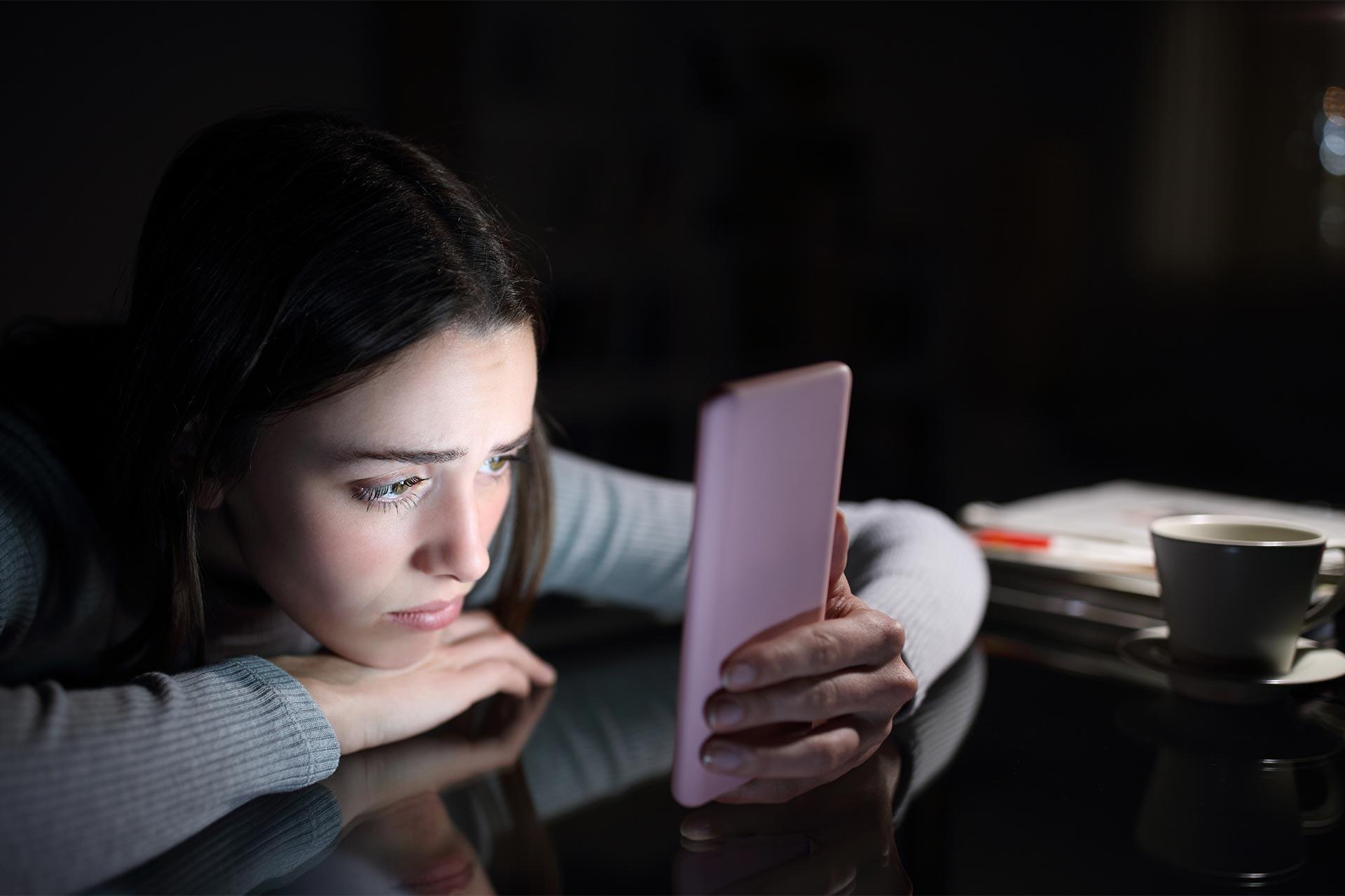 دختر جوان در تاریکی با چهره غمگین درحال تماشای نمایشگر گوشی است