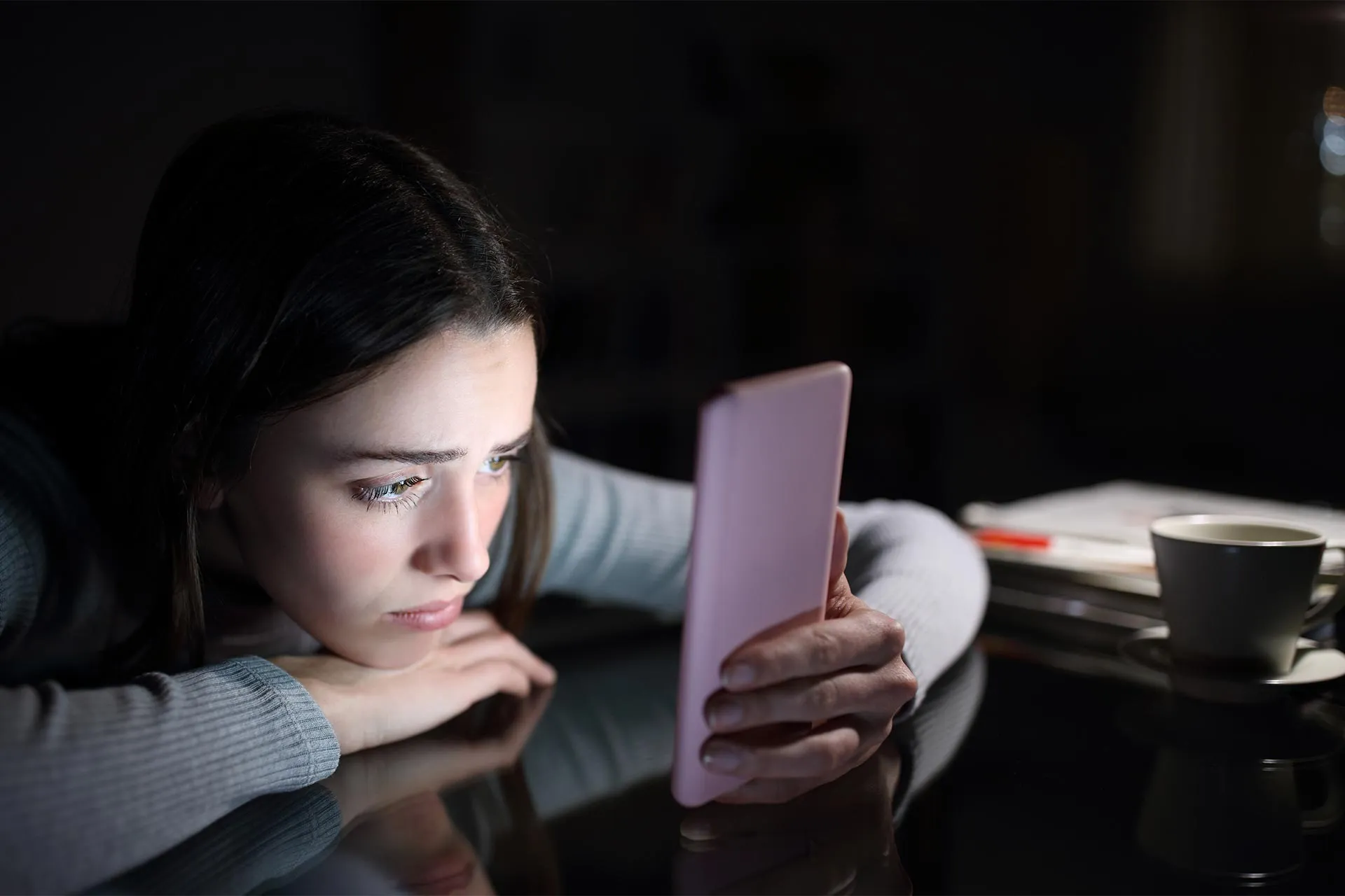 دختر جوان در تاریکی با چهره غمگین درحال تماشای نمایشگر گوشی است