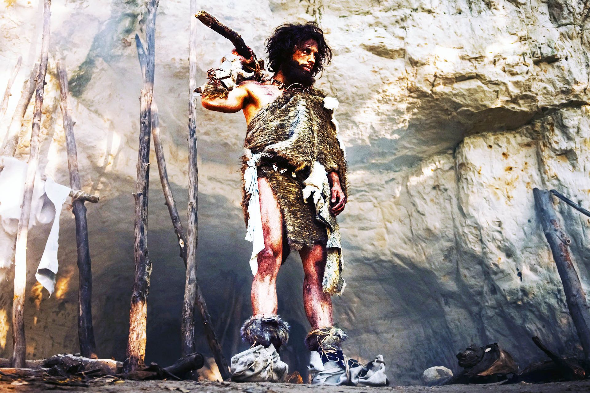 انسان غارنشین اولیه با پوست حیوان و نیزه نوک سنگی در دست، با لباس پشمی، در ورودی غار به اطراف جنگل پیشاتاریخی نگاه می‌کند.