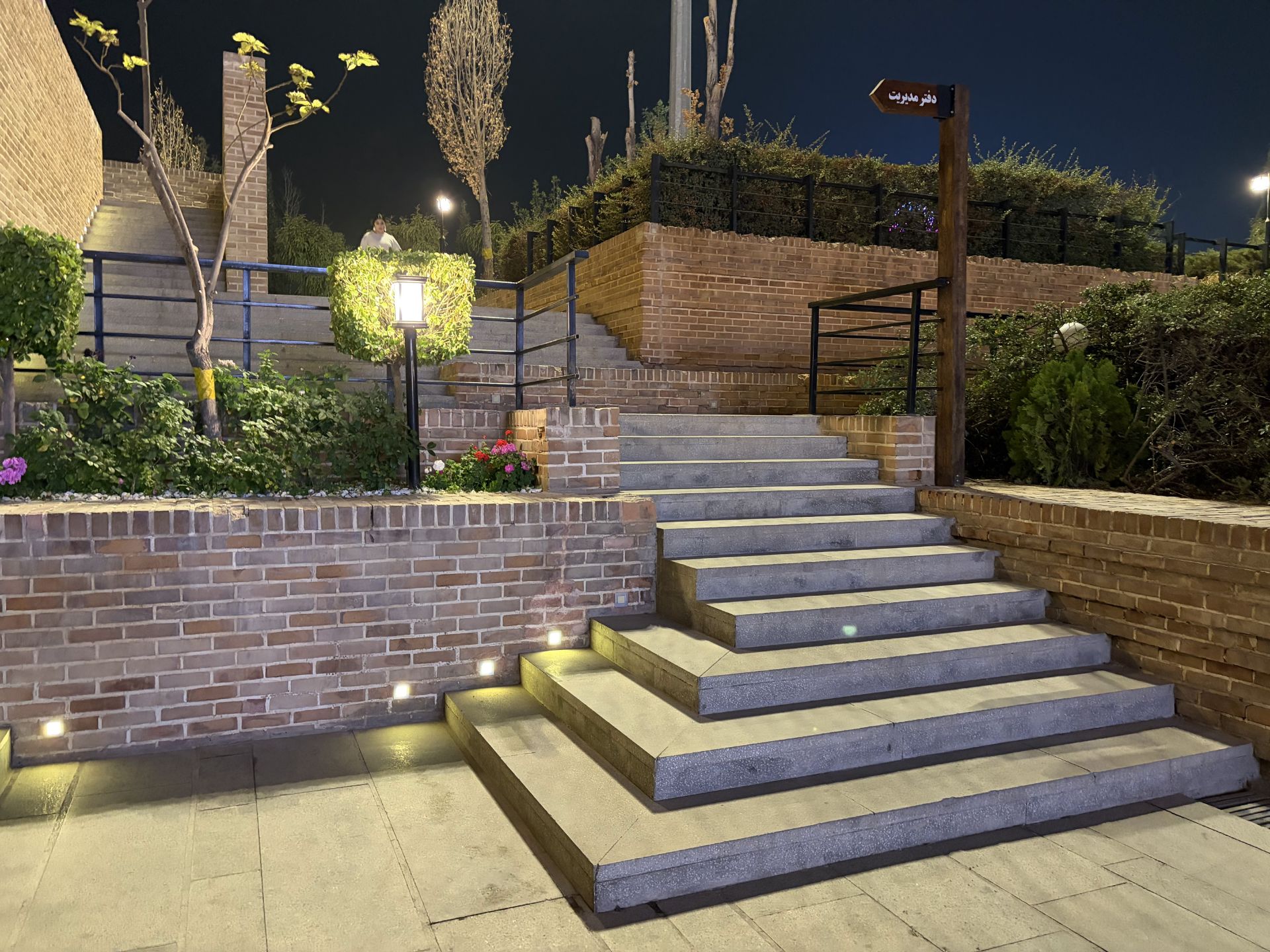 راه‌پله سنگی و دیوارهای آجری در فضای باز با بوته‌ها و چراغ‌های کوچک