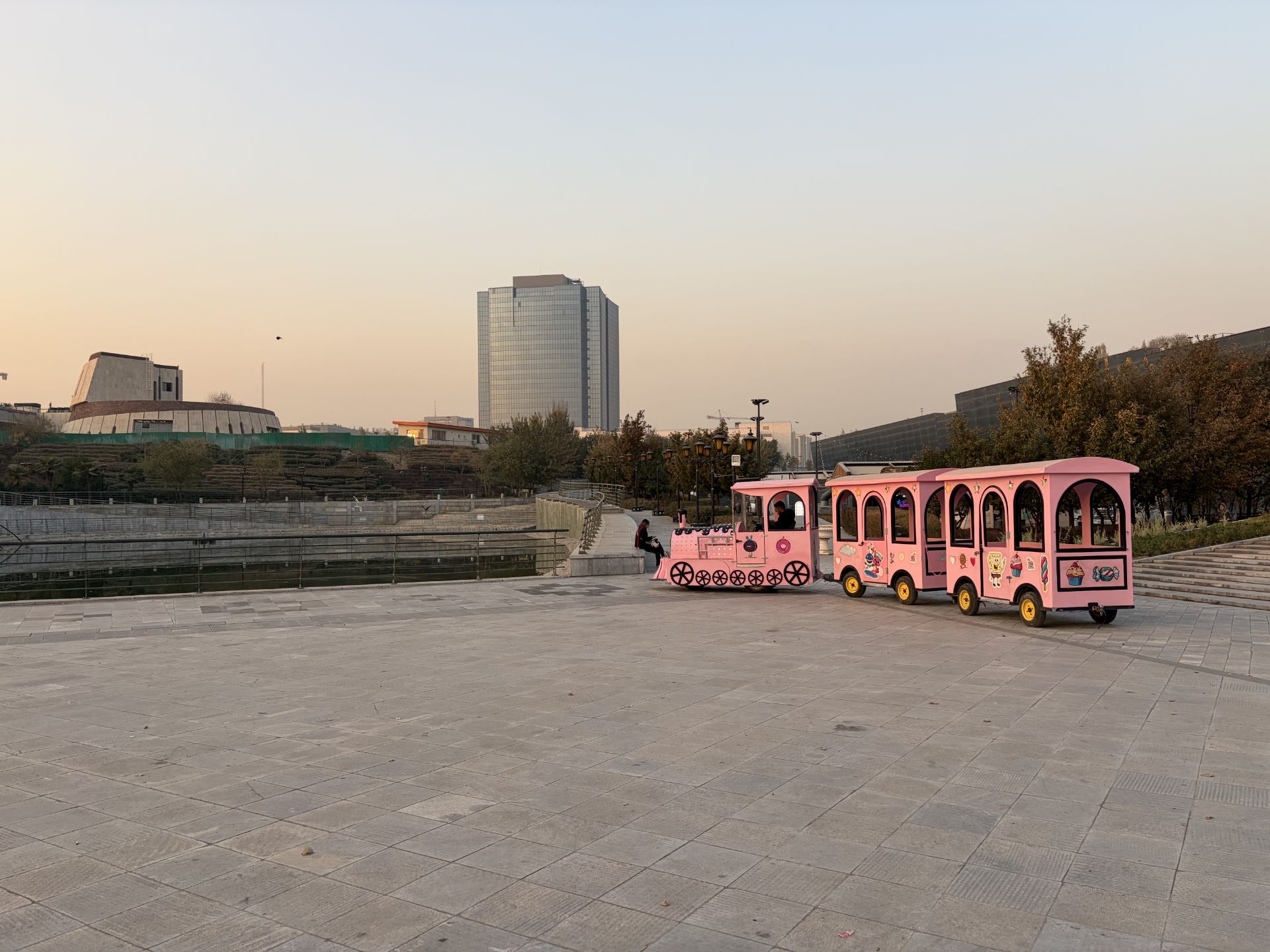 قطار تفریحی صورتی در محوطه سنگ‌فرش با ساختمان‌های بلند در پس‌زمینه هنگام غروب