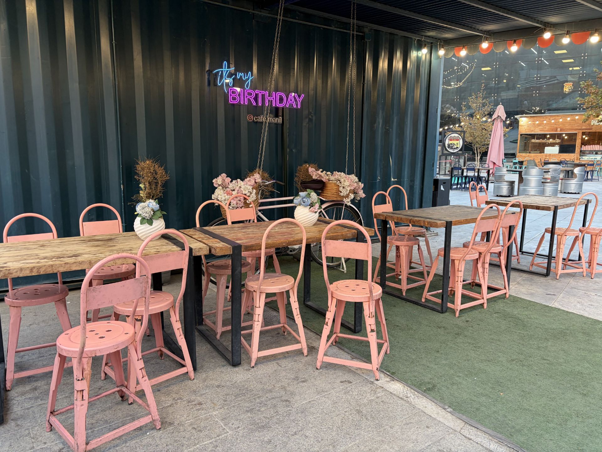 میز و صندلی‌های صورتی کافه در فضای باز با تابلوی نئون «It’s my birthday»
