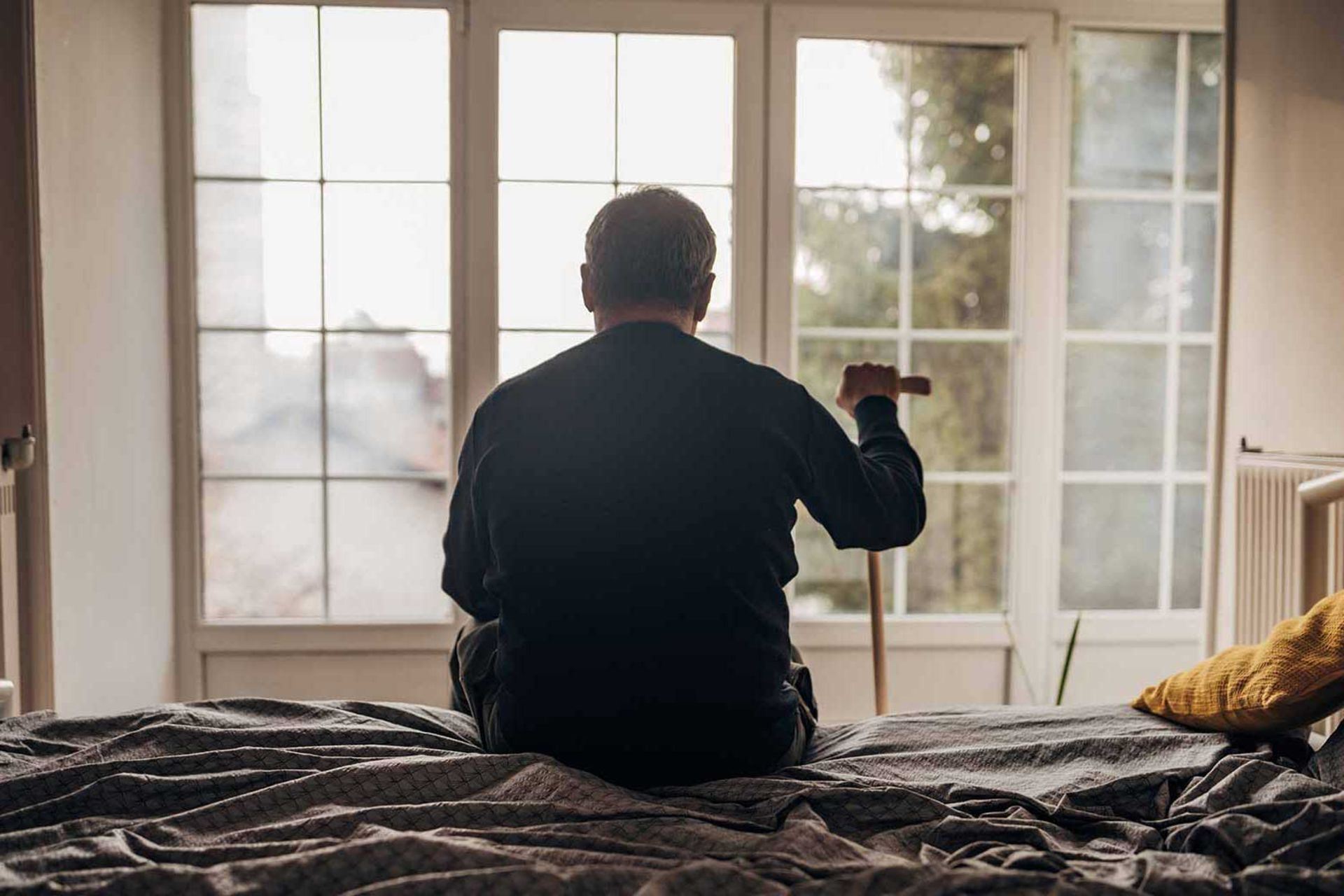 مرد مسن تنها پشت به دوربین و عصا به دست روی تخت رو به پنجره نشسته است