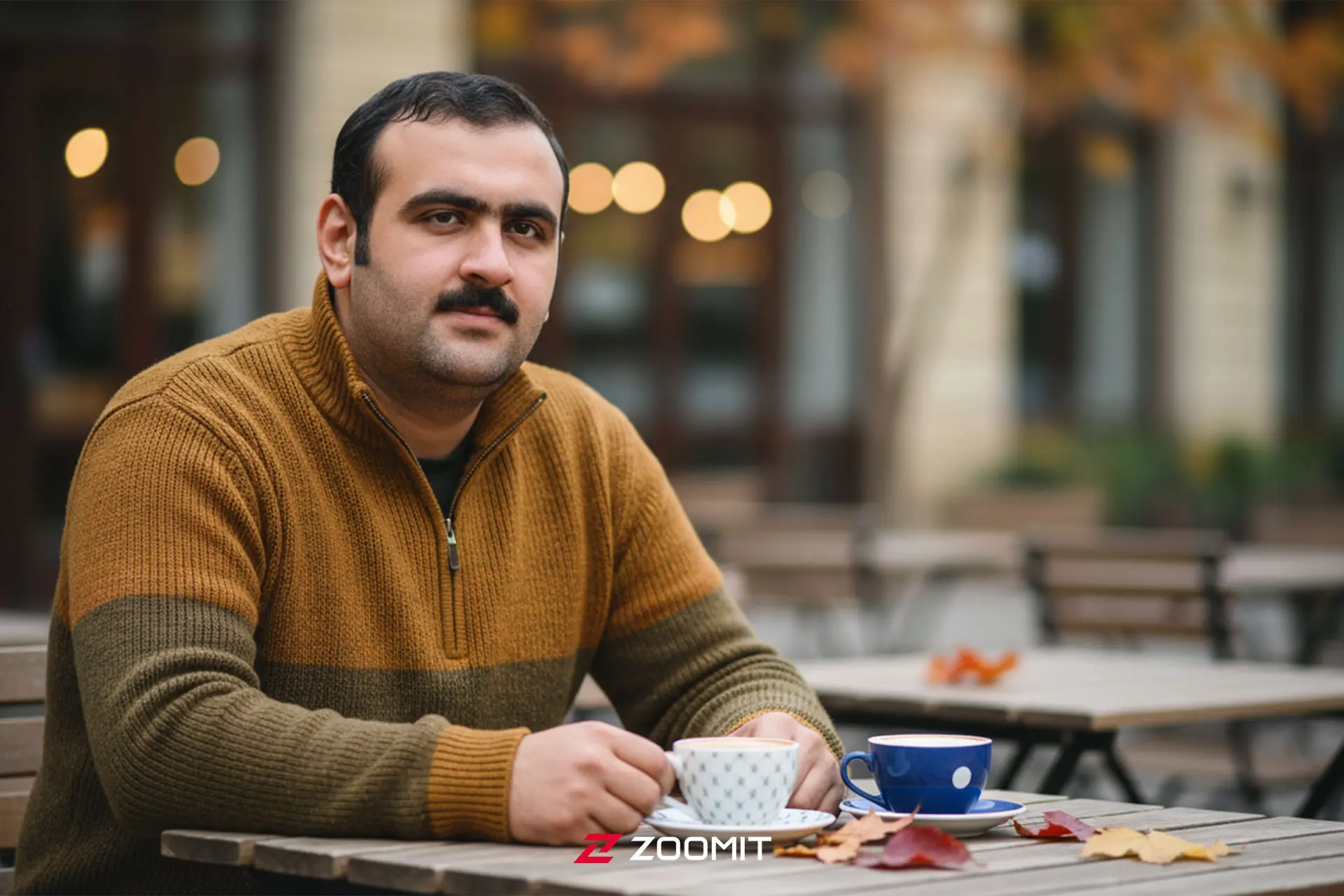 مردی با پلیور قهوه‌ای در کافه روباز پاییزی نشسته