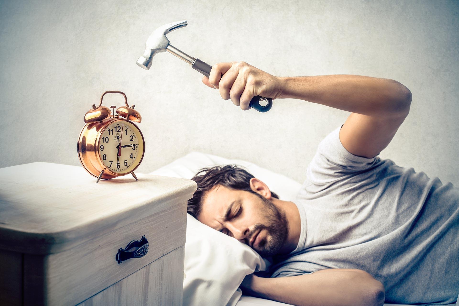 مرد جوان روی تخت خوابیده و در تلاش برای ضربه‌زدن به ساعت زنگ‌دار با چکش
