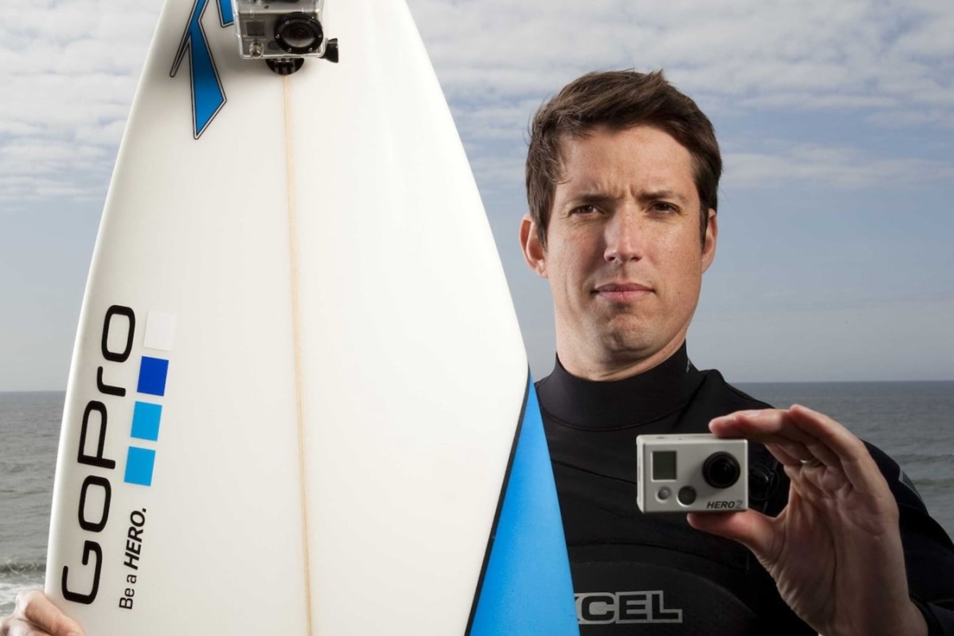 یک مرد جوان و دوربین گوپرو و تخته‌ی موج‌سواری در دست