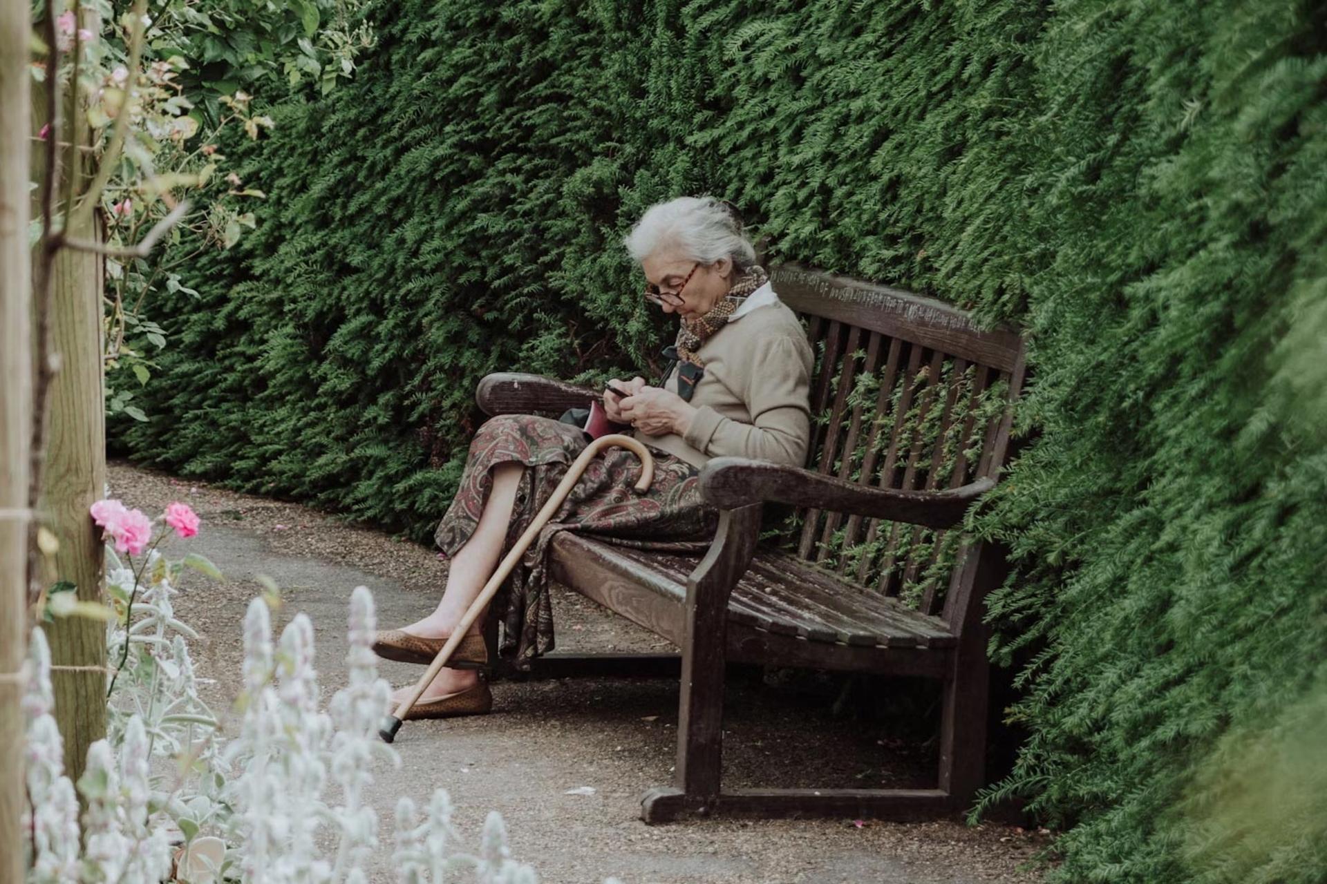 خانم سالمندی روی یک نیمکت در پارک نشسته و با گوشی کار می‌کند