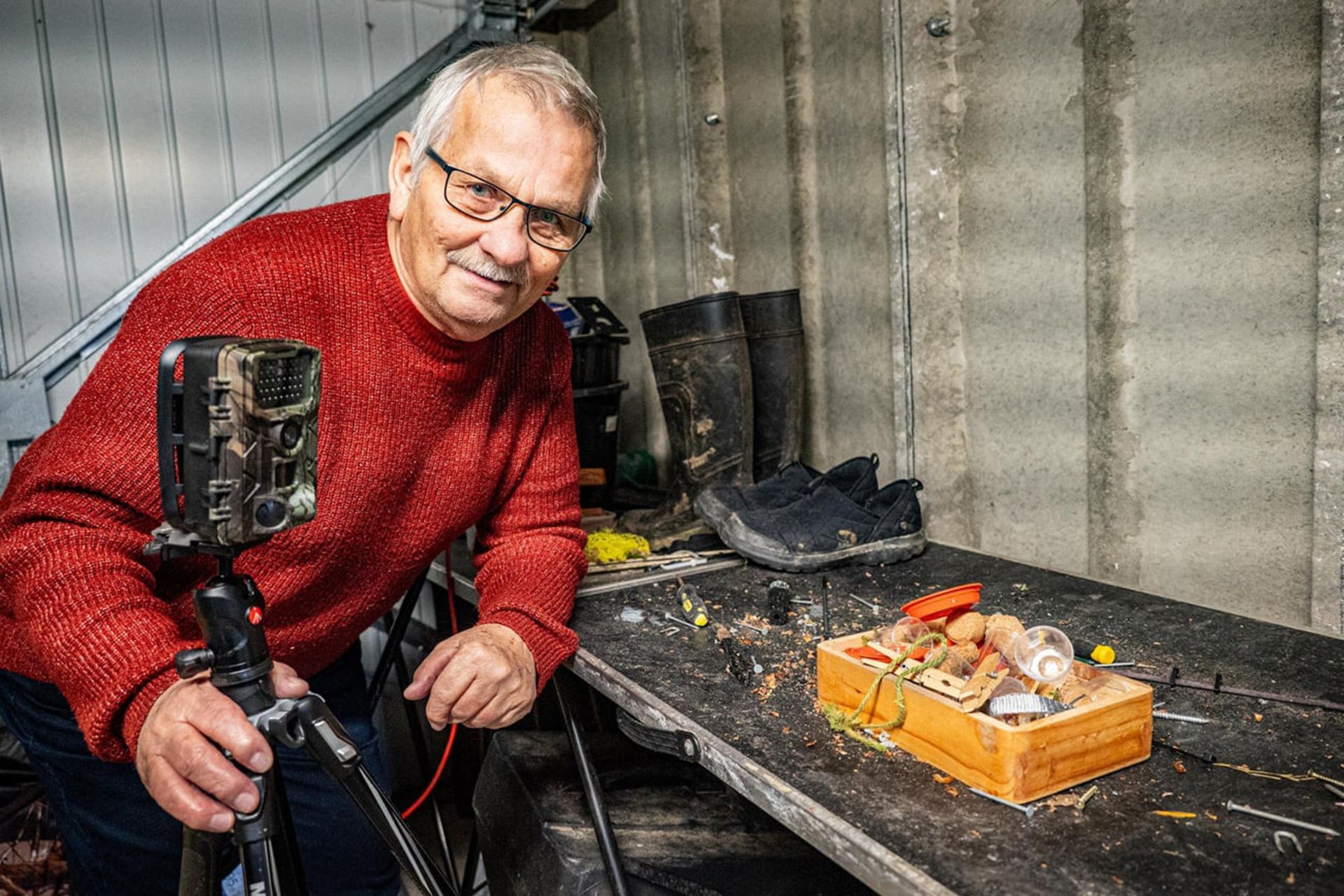 پیرمردی با لباس قرمز که در کنار میز کارش قرار گرفته و پایه دوربین در دست دارد