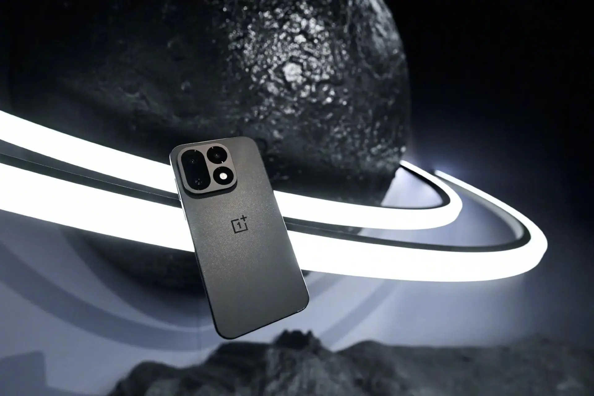 گوشی هوشمند وان‌پلاس ۱۶ (OnePlus 16) با بدنه خاکستری در کنار یک کره سیاه بزرگ