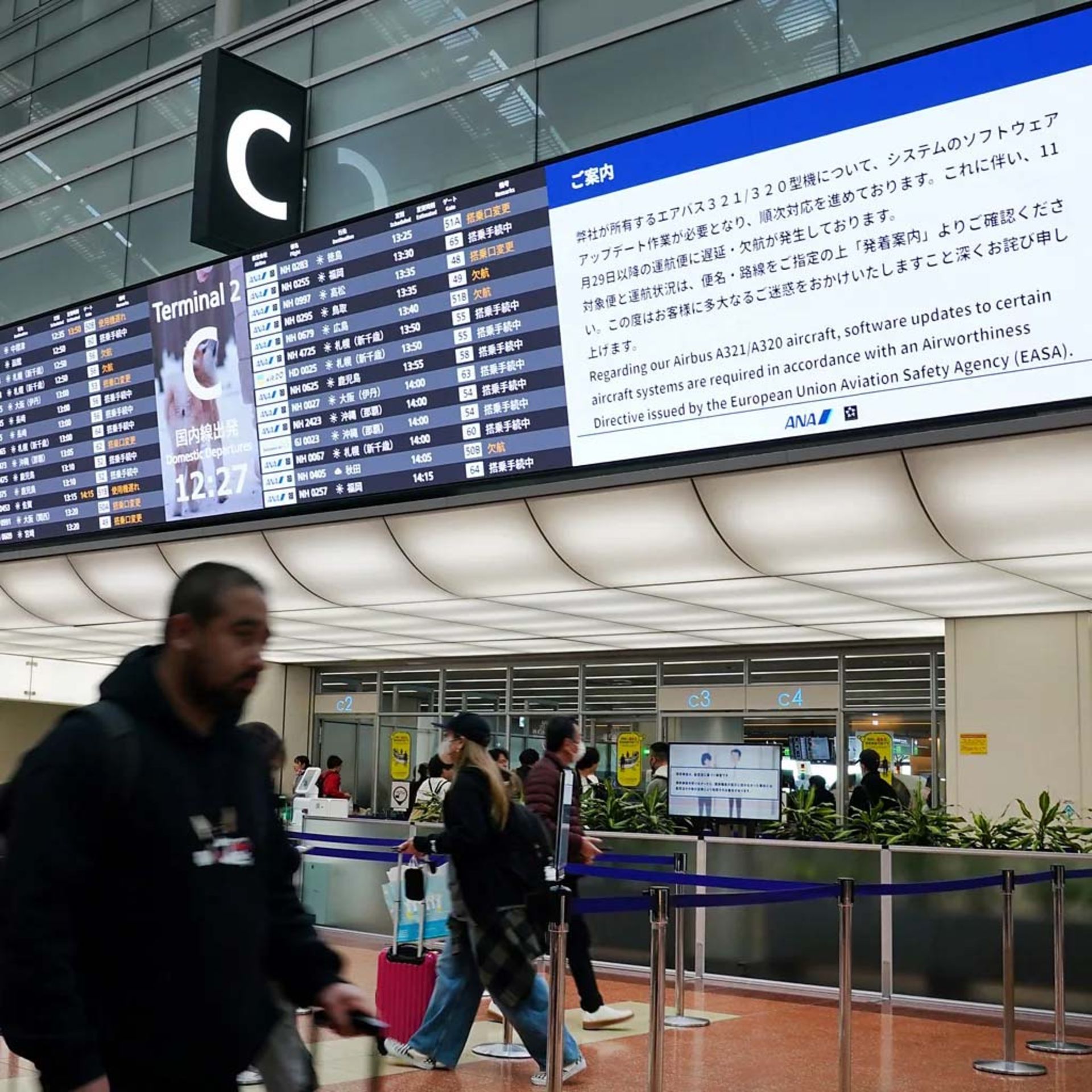 افراد زیر تابلوی اعلانات سالن فرودگاه راه می‌روند 