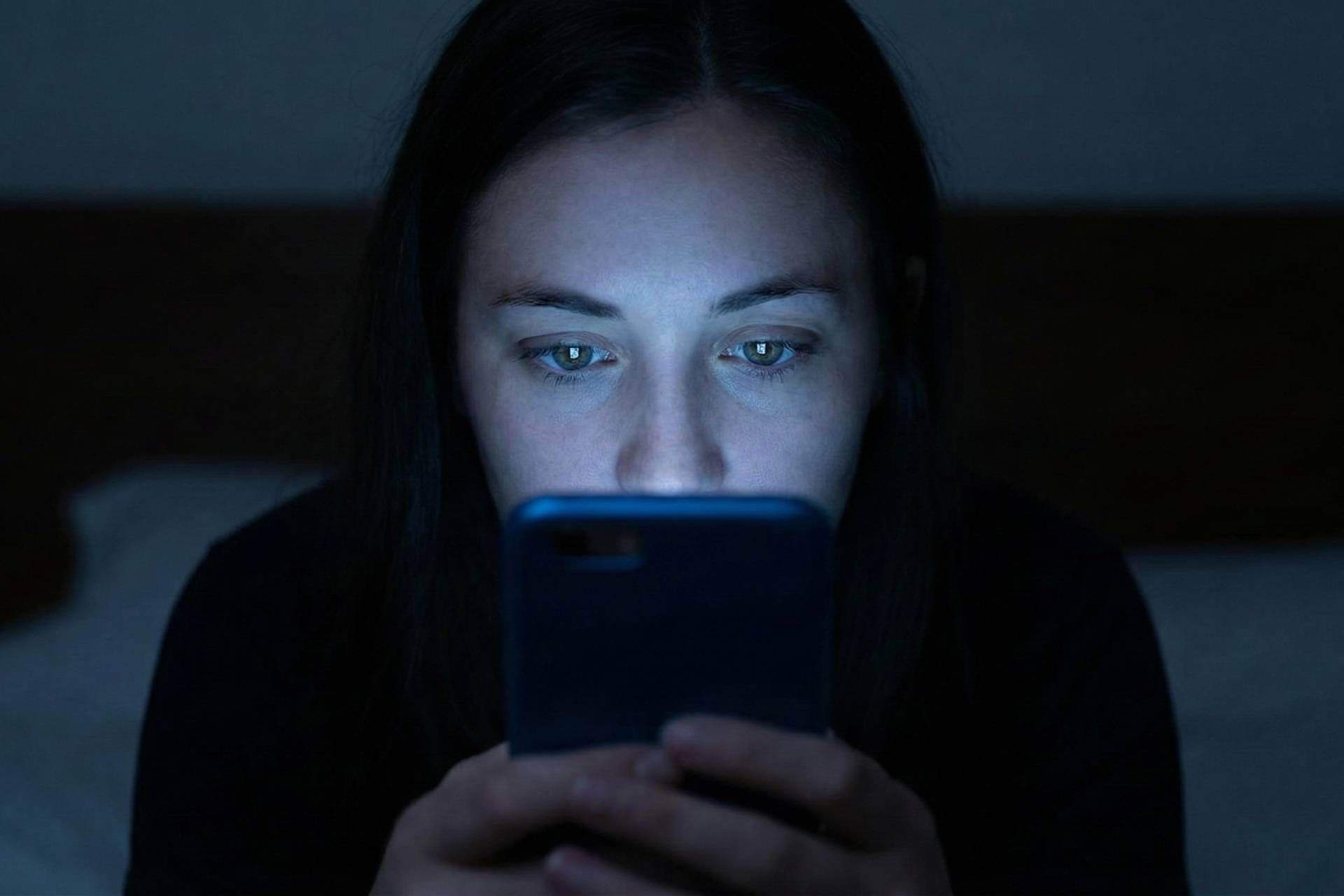 چهره فردی که تنها با نور آبی گوشی هوشمندش در یک اتاق تاریک روشن شده است.