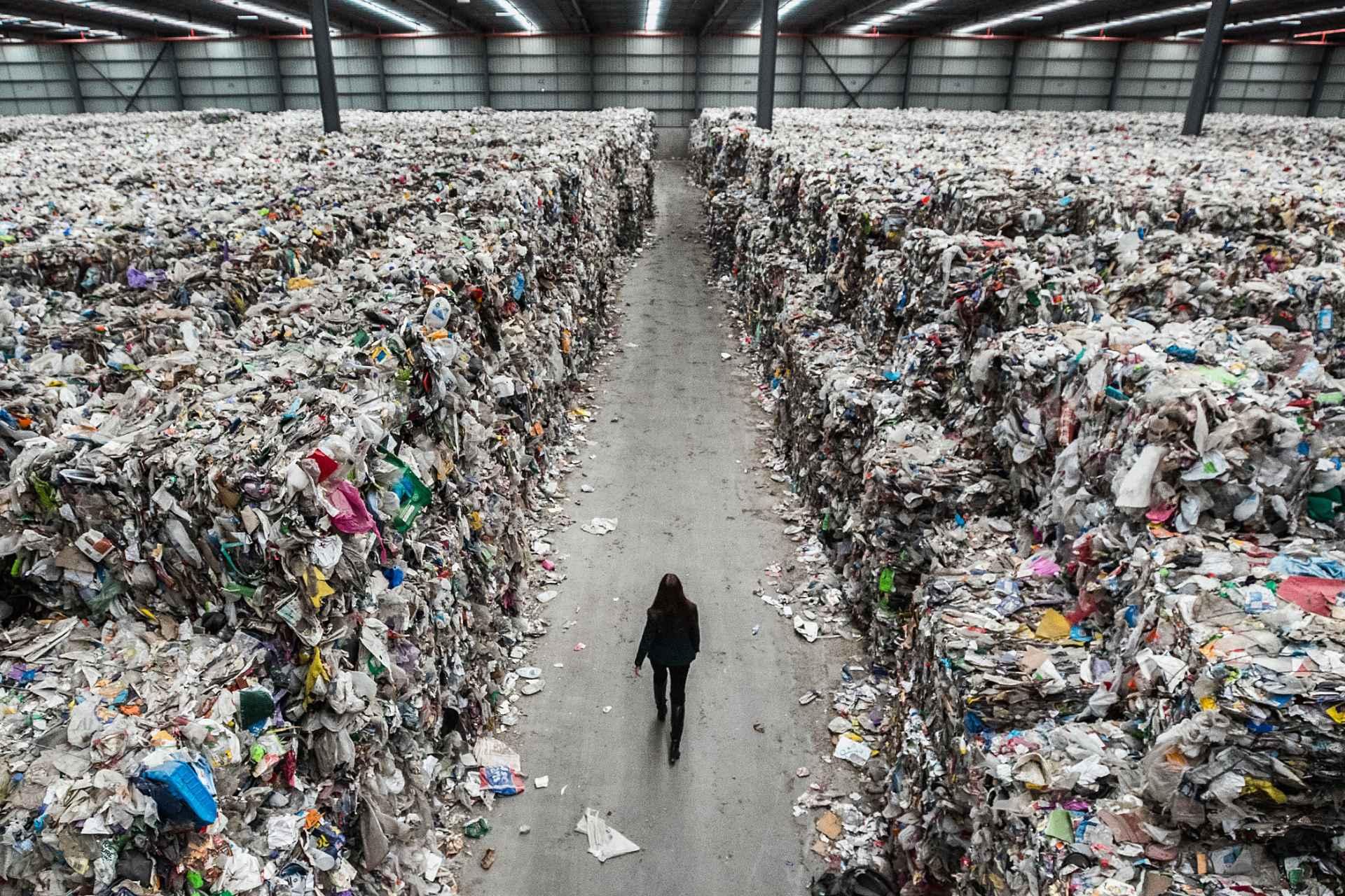 فردی در راهروی بین کوه‌های زباله در سوله‌ای بزرگ قدم می‌زند.