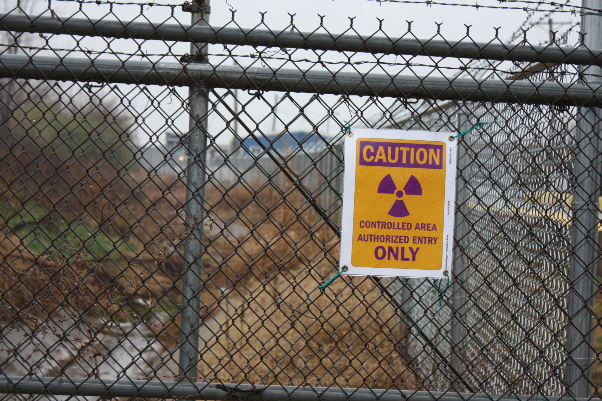 تابلوی هشدار رادیواکتیو پشت حصار فلزی در منطقه‌ای کنترل‌شده با دسترسی محدود در محوطه‌ای آلوده به پسماند هسته‌ای