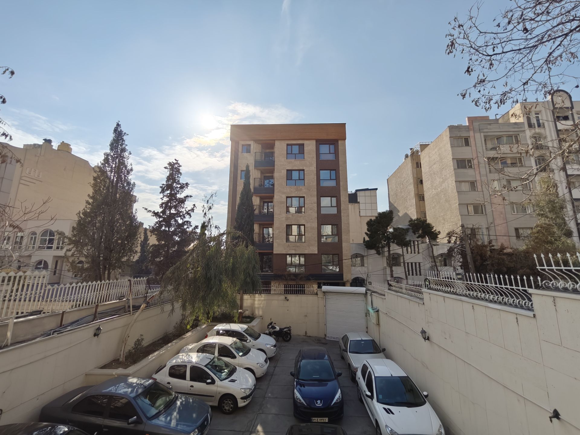 ساختمان در روبرو با پس‌زمینه آسمان آفتابی و ماشین‌ها در حیاط 