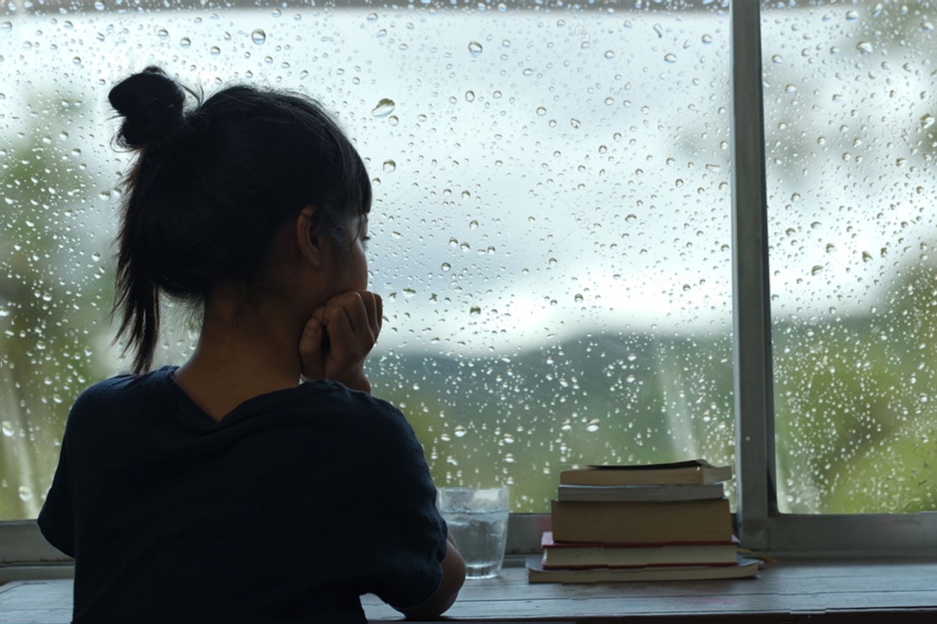 دختر نوجوان ناراحت با تیشرت سرمه‌ای و موهای گوجه‌ای از پنجره به باران نگاه می‌کند