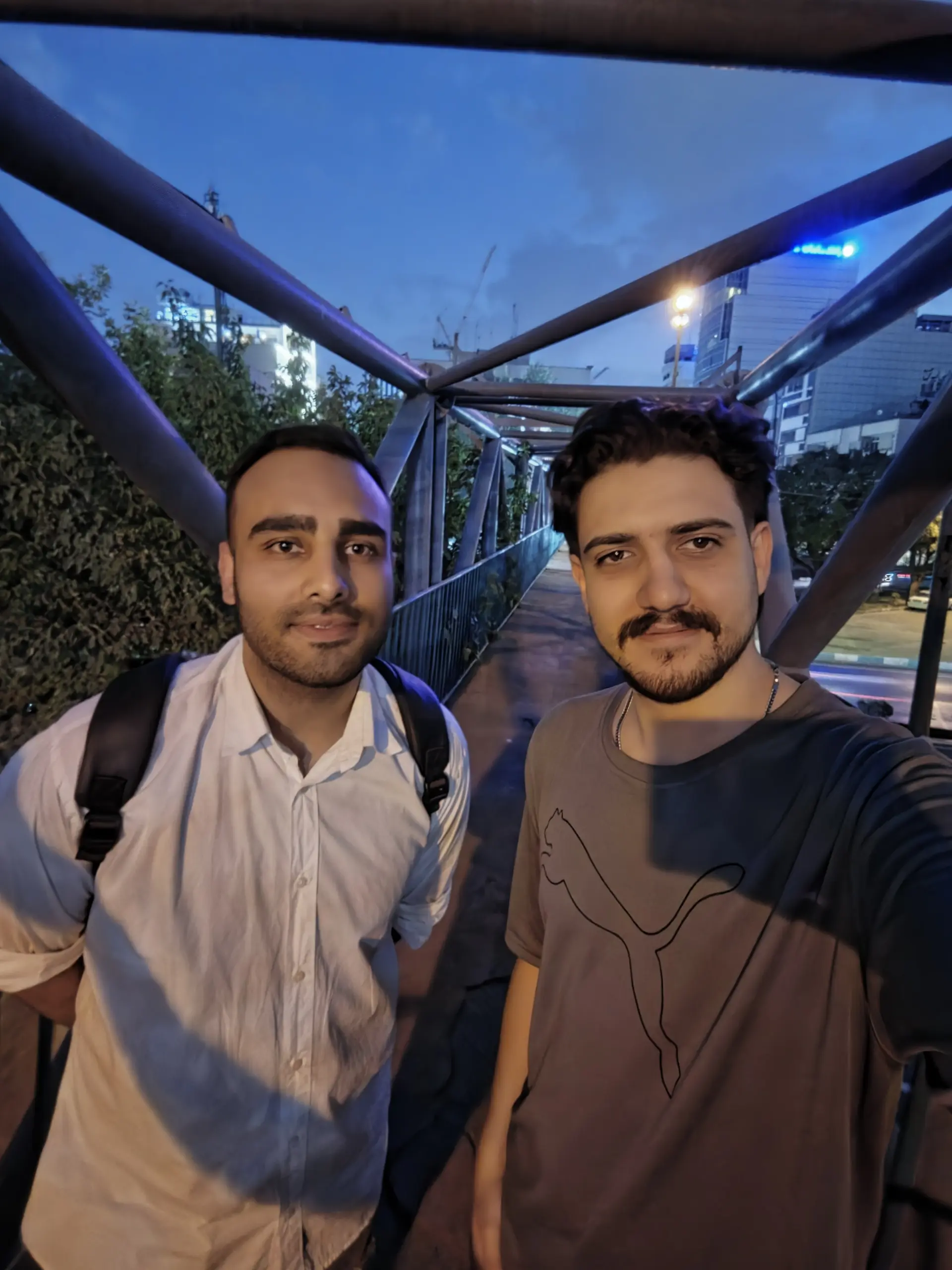 دو مرد جوان در یک مسیر پیادهرو با پسزمینه ساختمانهای شهری در شب
