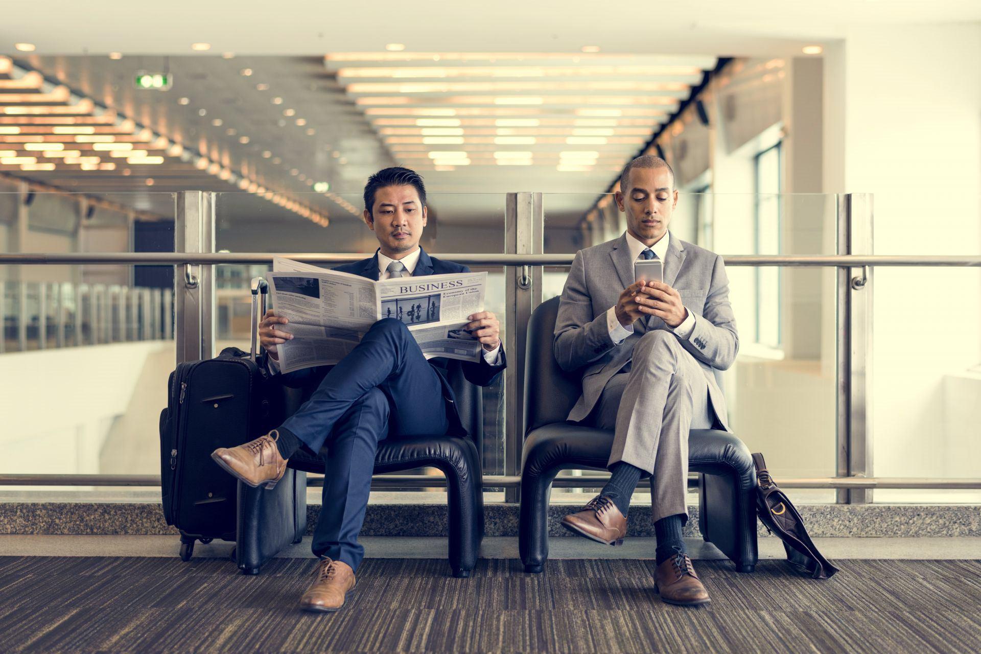 دو مرد که پاهایشان را روی هم انداخته‌اند درحال خواندن روزنامه و چک‌کردن موبایل