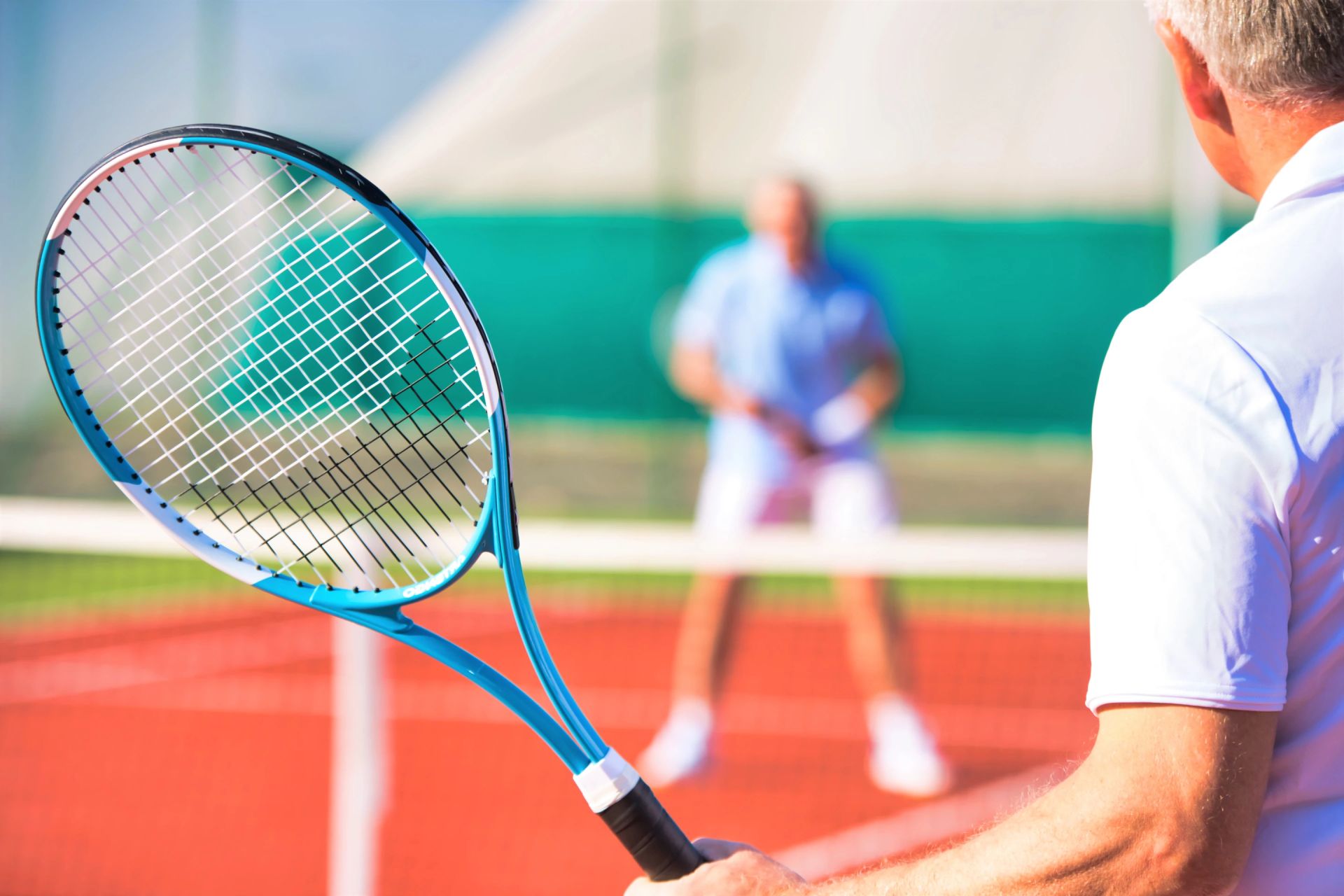 نمای نزدیک راکت تنیس در دست مردی با لباس سفید در حال بازی با مرد دیگری با سایه محو در پس زمینه