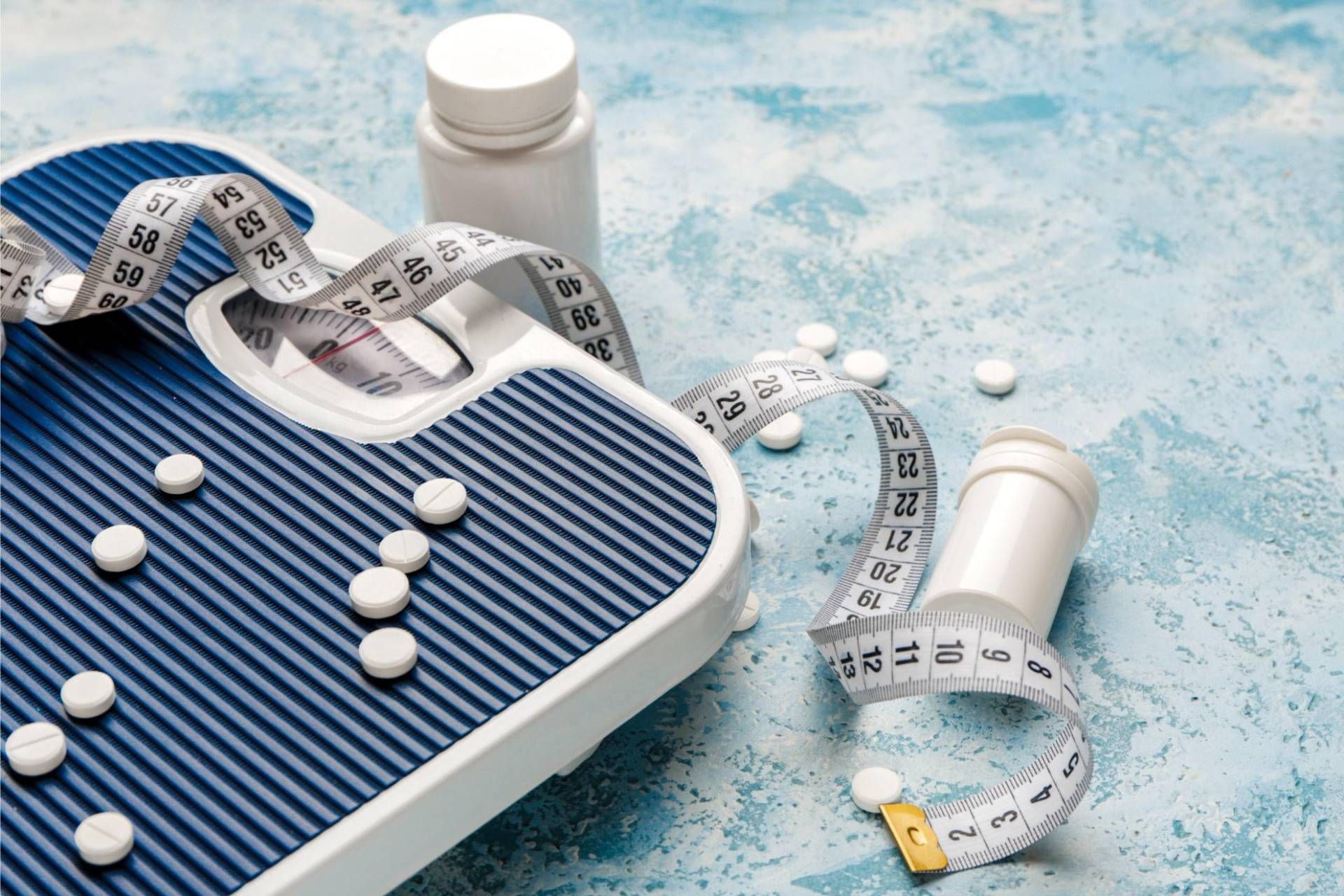 ترازو وزن‌کشی با نوار اندازه‌گیری پیچ‌خورده روی آن و چند قرص سفید و قوطی دارو در اطراف.