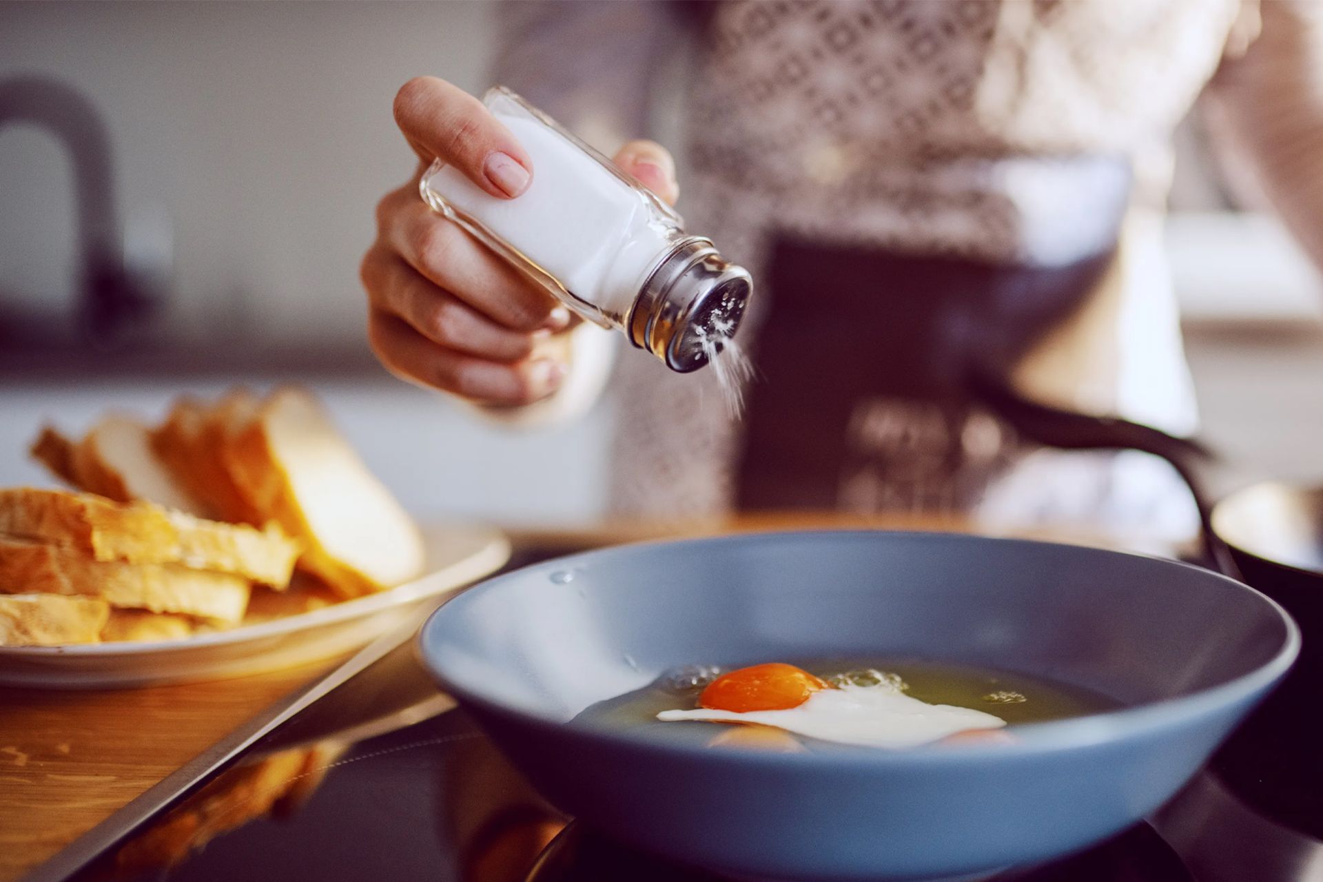 نمای نزدیک از زنی کنار اجاق گاز آشپزخانه در حال افزودن نمک به تخم‌مرغ توی ماهیتابه
