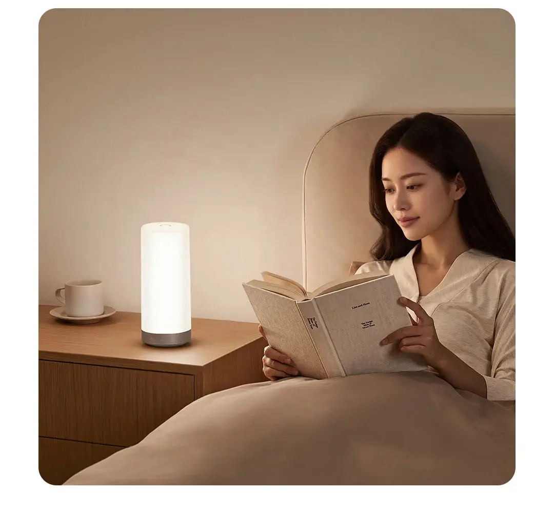 چراغ‌خواب هوشمند شیائومی (Xiaomi) روی میز کنار تخت در اتاق خواب کنار یک زن در حال مطالعه